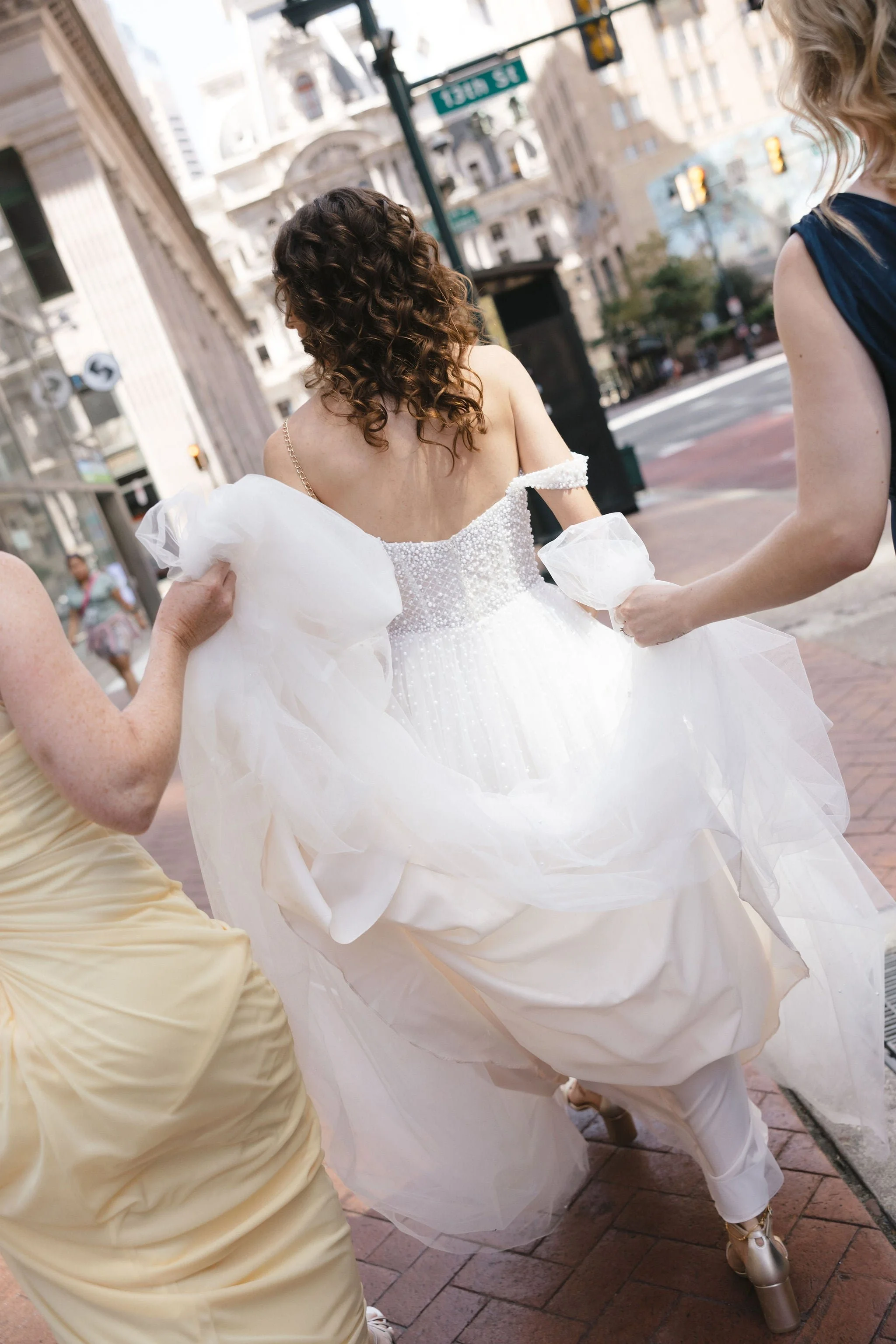 A bride in a white wedding dress with curly hair, being assisted by two women on a city street with tall buildings and traffic lights visible in the background.
