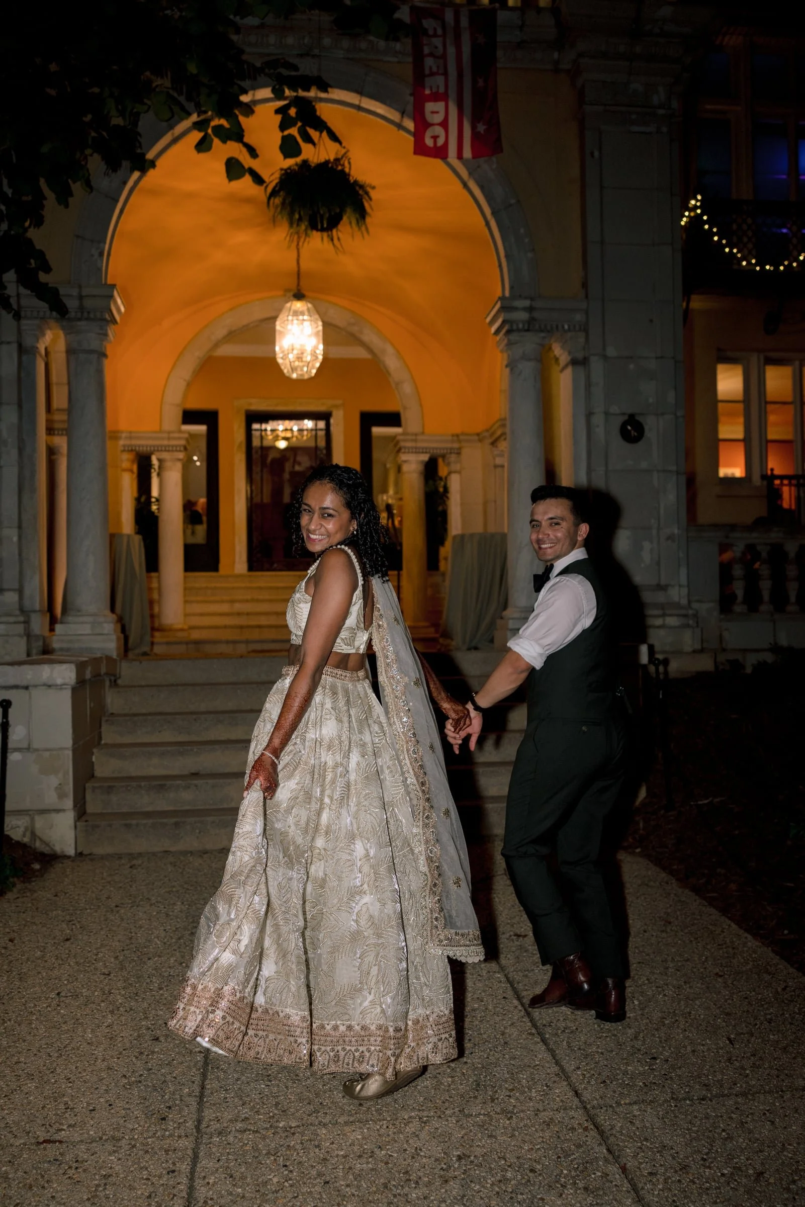 A smiling woman in a traditional Indian dress stands in front of a building with columns and arches. She holds hands with a man wearing a black vest and white shirt. The scene is lit with warm lighting, and there is a chandelier hanging from the ceil