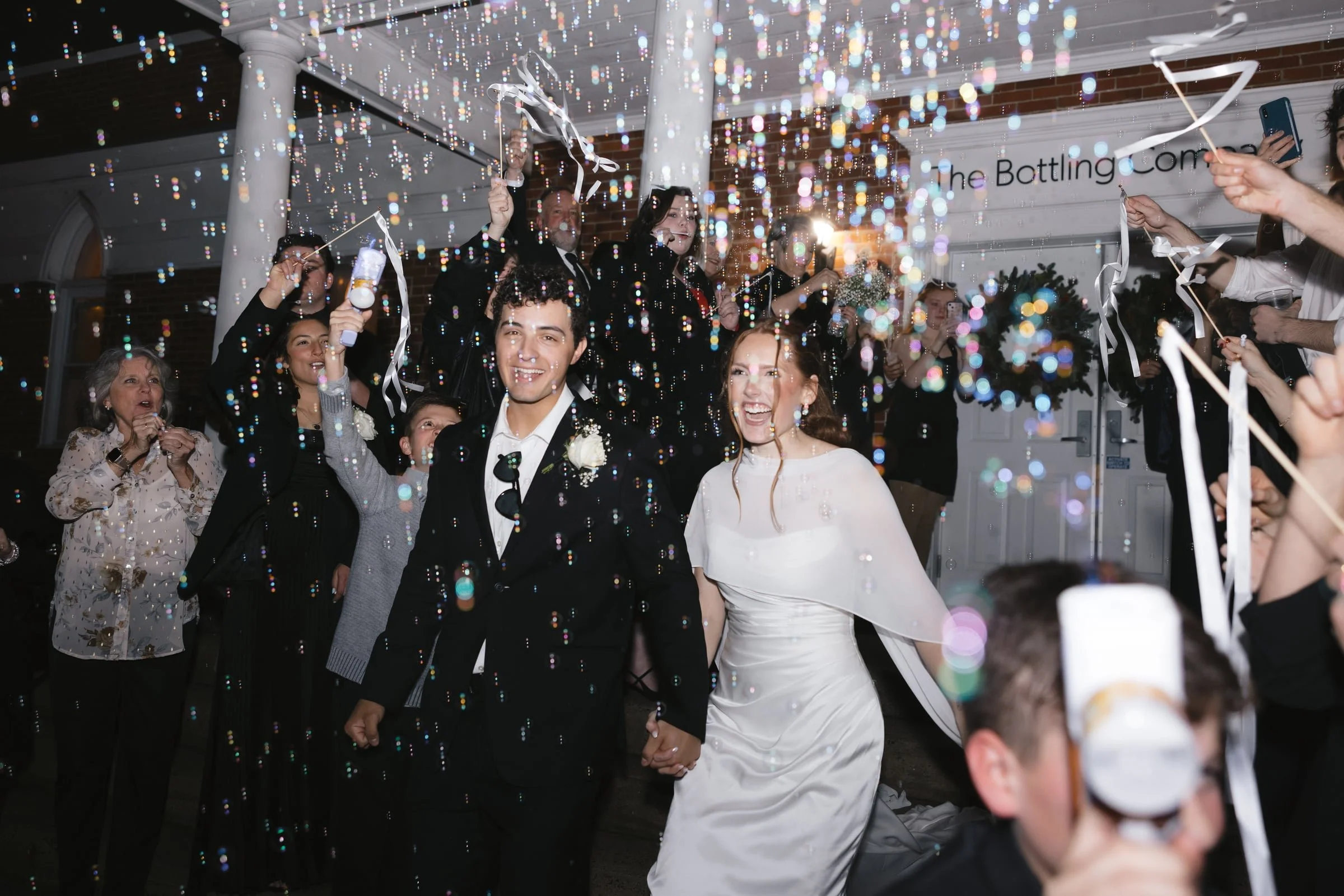 A celebration scene with a bride and groom holding hands in front of a group of people, surrounded by colorful confetti and party decorations at a party venue.