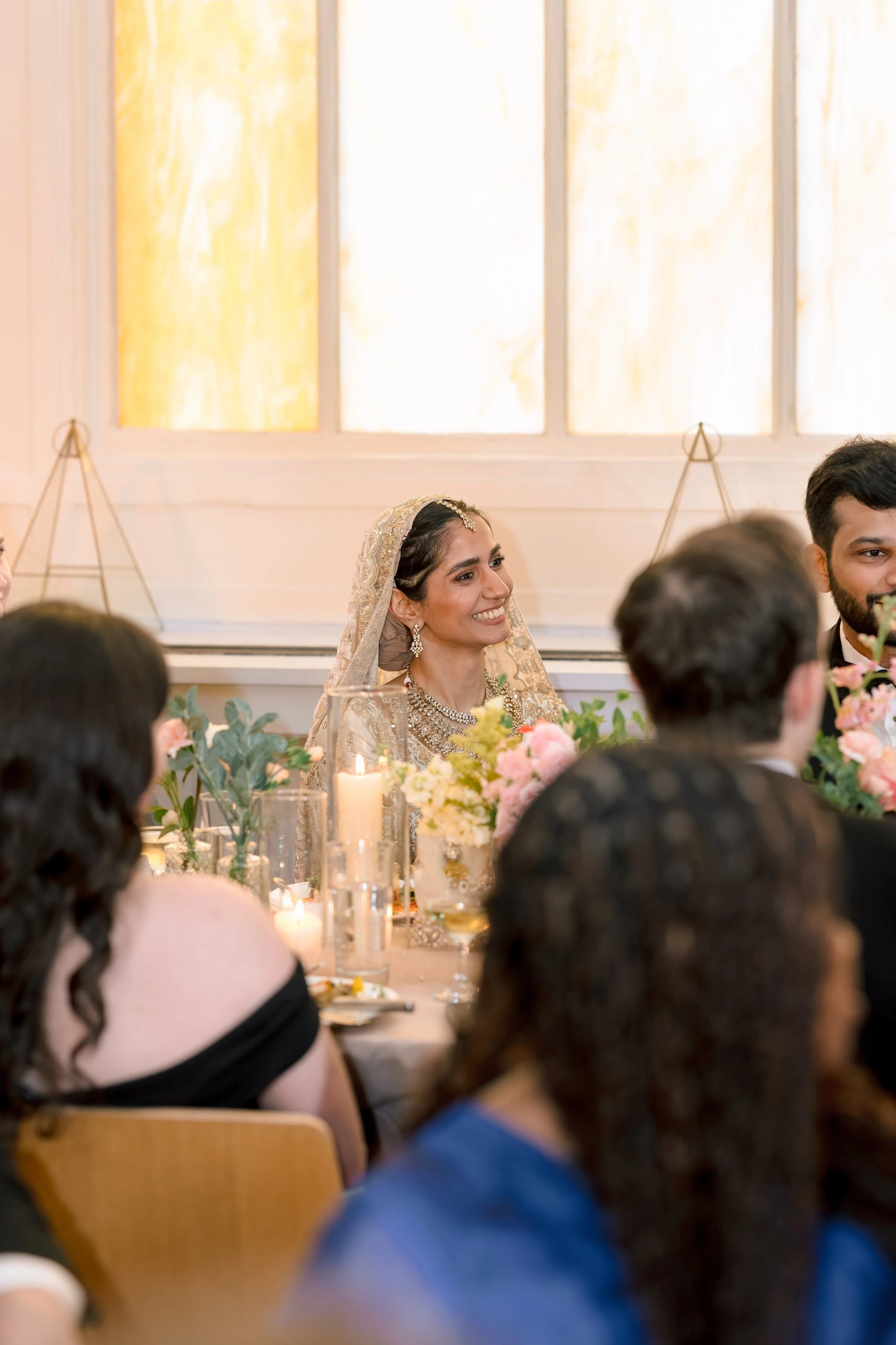 People gathered at a wedding reception with a bride in traditional attire smiling, surrounded by floral decorations and guests seated at a table with candles.