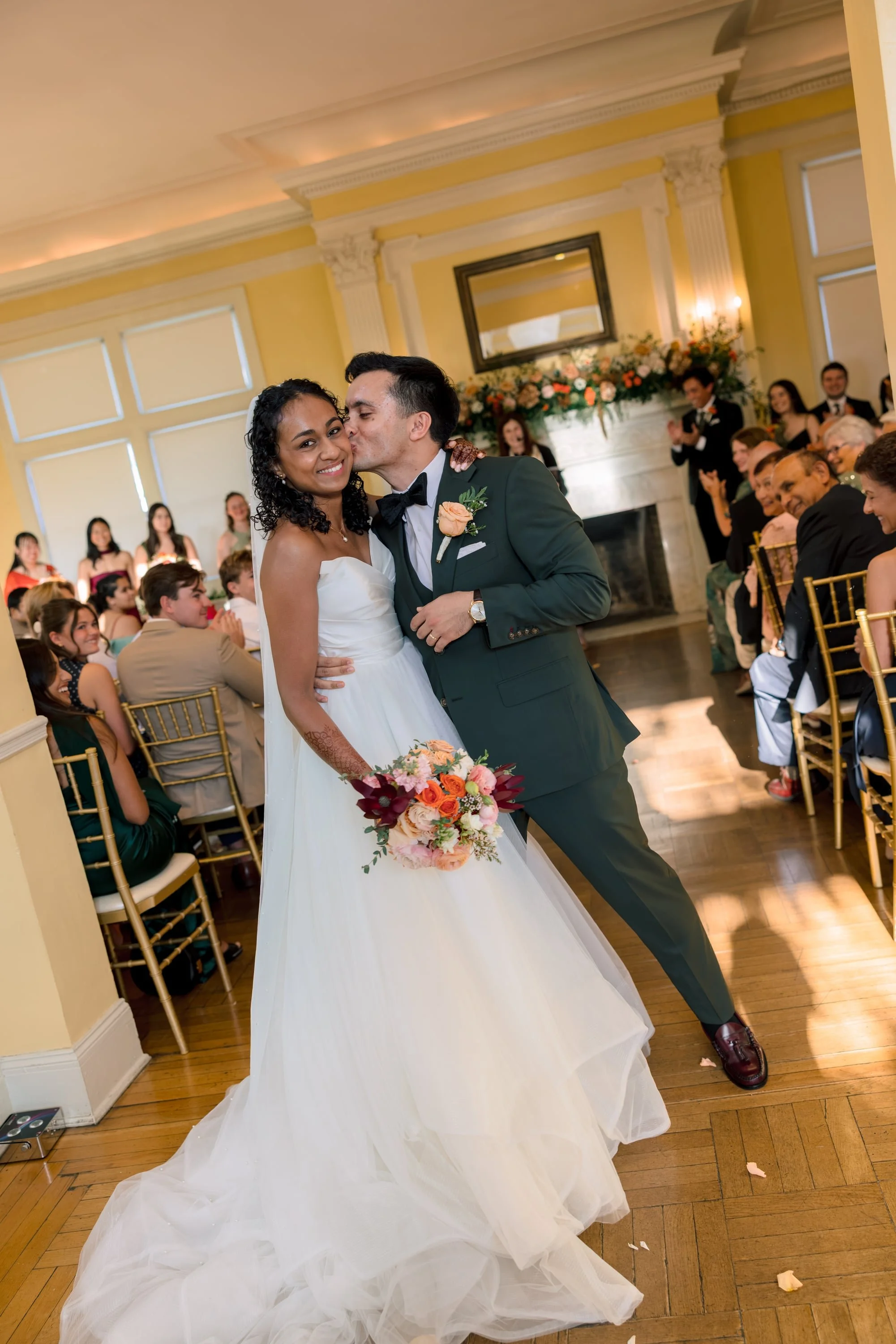 A bride and groom sharing a kiss at their wedding ceremony, with wedding guests sitting on gold chairs in the background, floral decorations on the fireplace, and the bride holding a bouquet of colorful flowers.