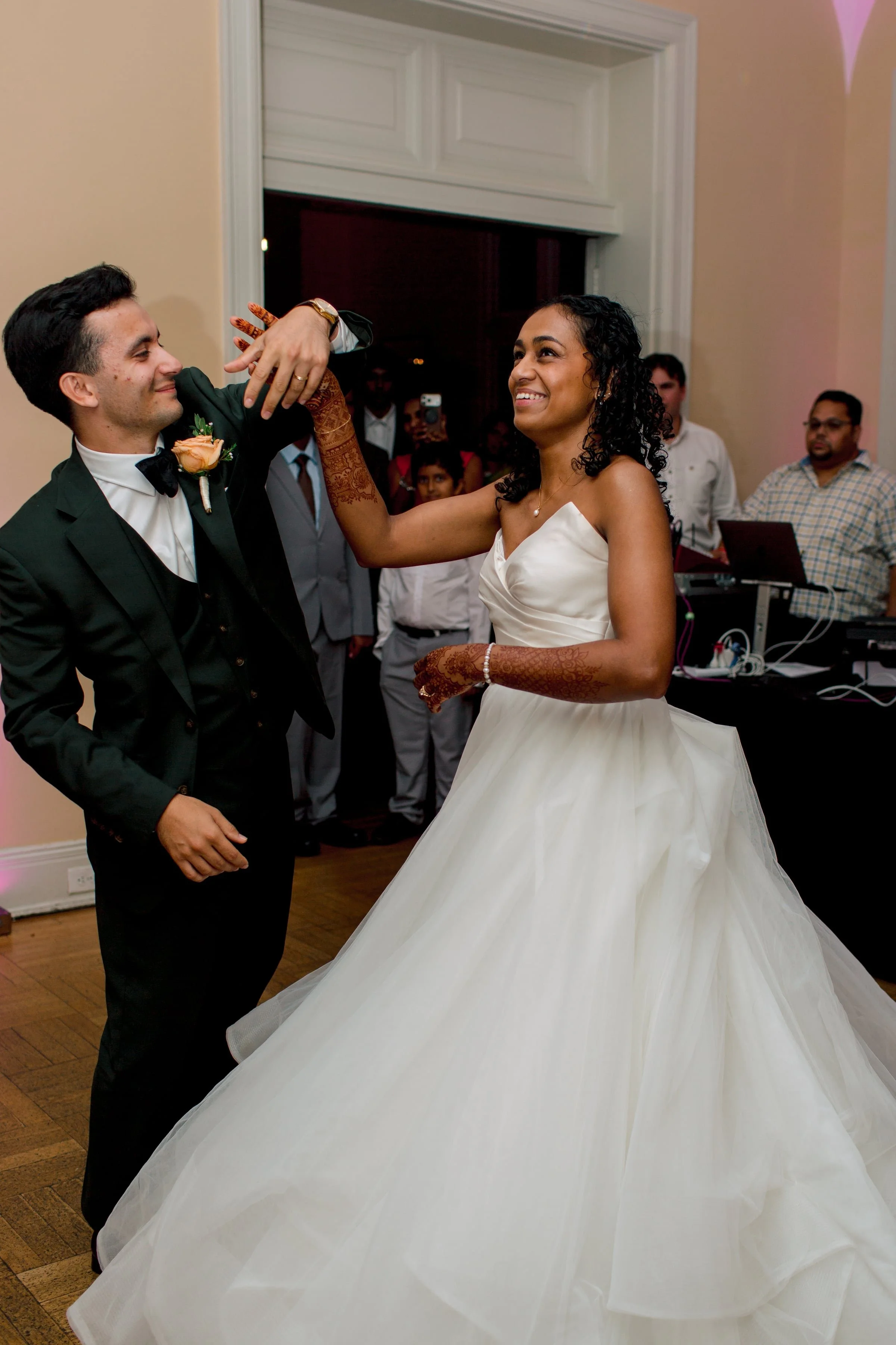 A bride and groom dancing at their wedding reception, with the bride in a strapless white wedding gown, and the groom in a black tuxedo, surrounded by guests.
