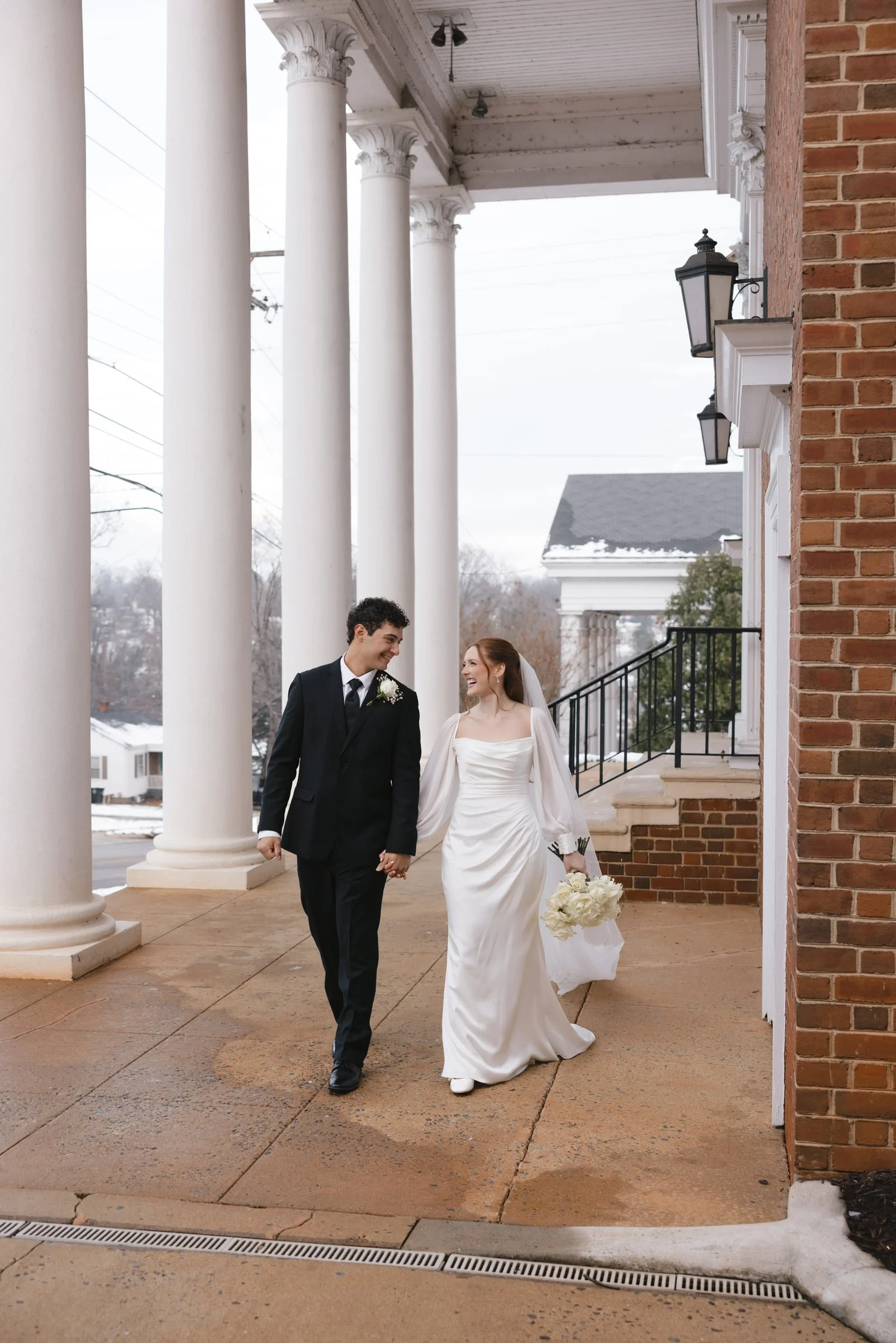 A newly married couple walking hand in hand outside a church, smiling and looking at each other, surrounded by white columns and brick walls, with snowy ground and a gray sky in the background.