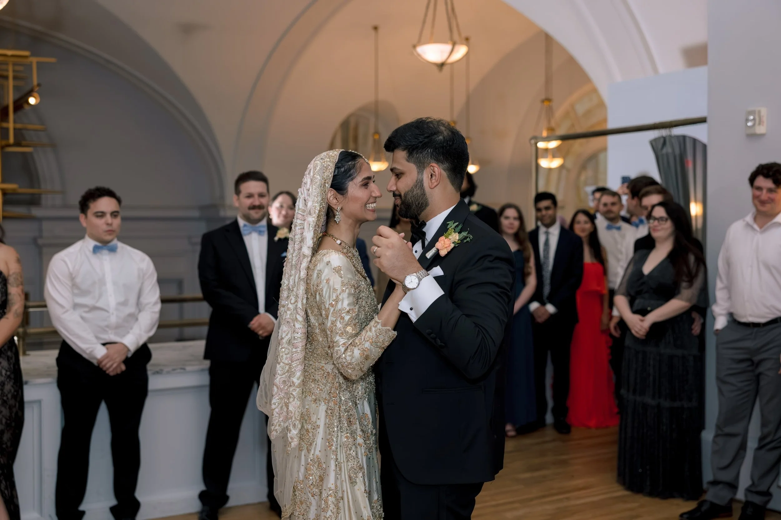 A bride and groom are dancing closely, smiling at each other, during their wedding reception in an elegant, well-lit hall with arched ceilings. Guests are watching and smiling in the background.