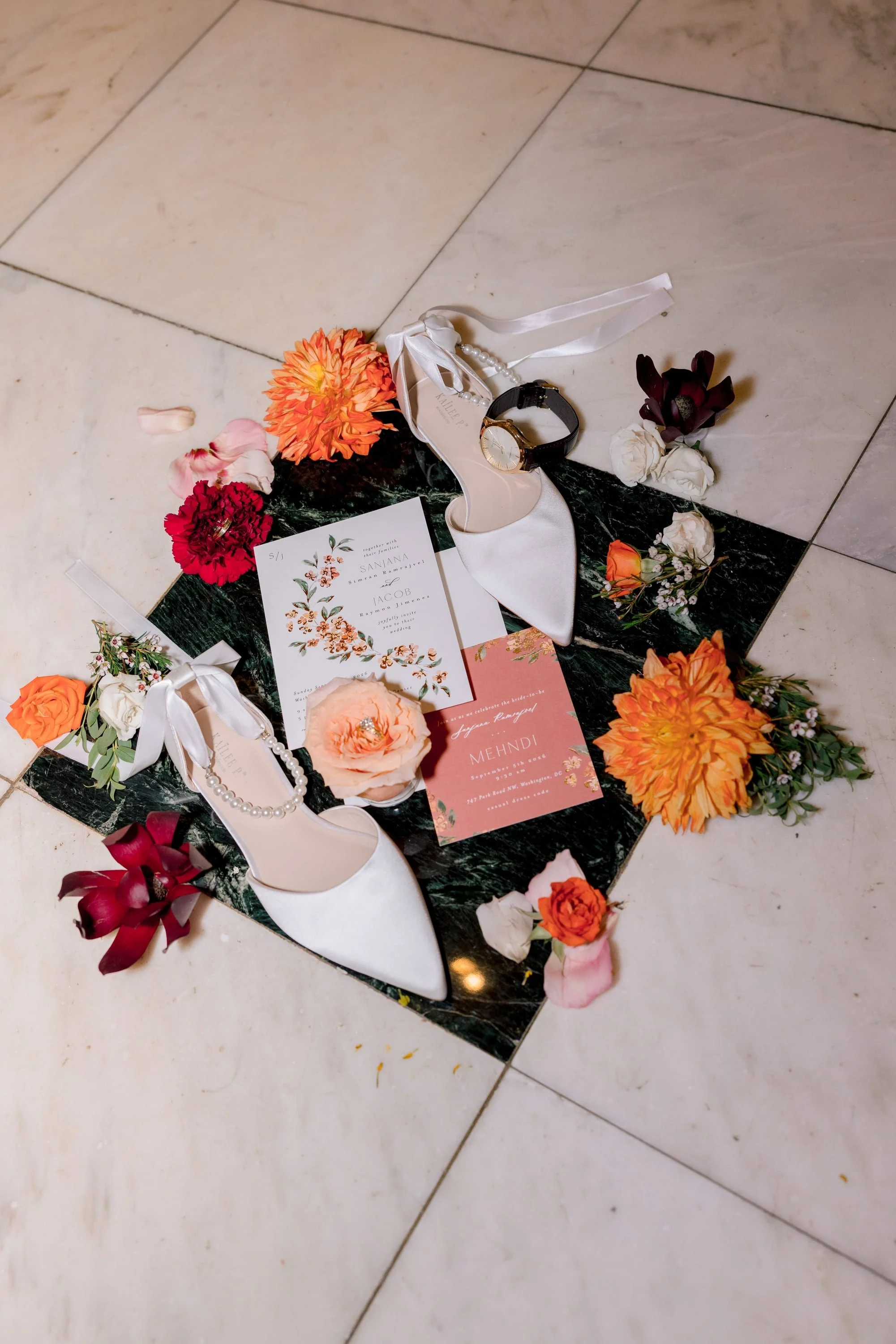 Wedding invitation, white high-heeled shoes, wristwatch, and various flowers arranged on black marble and white tiled floor.