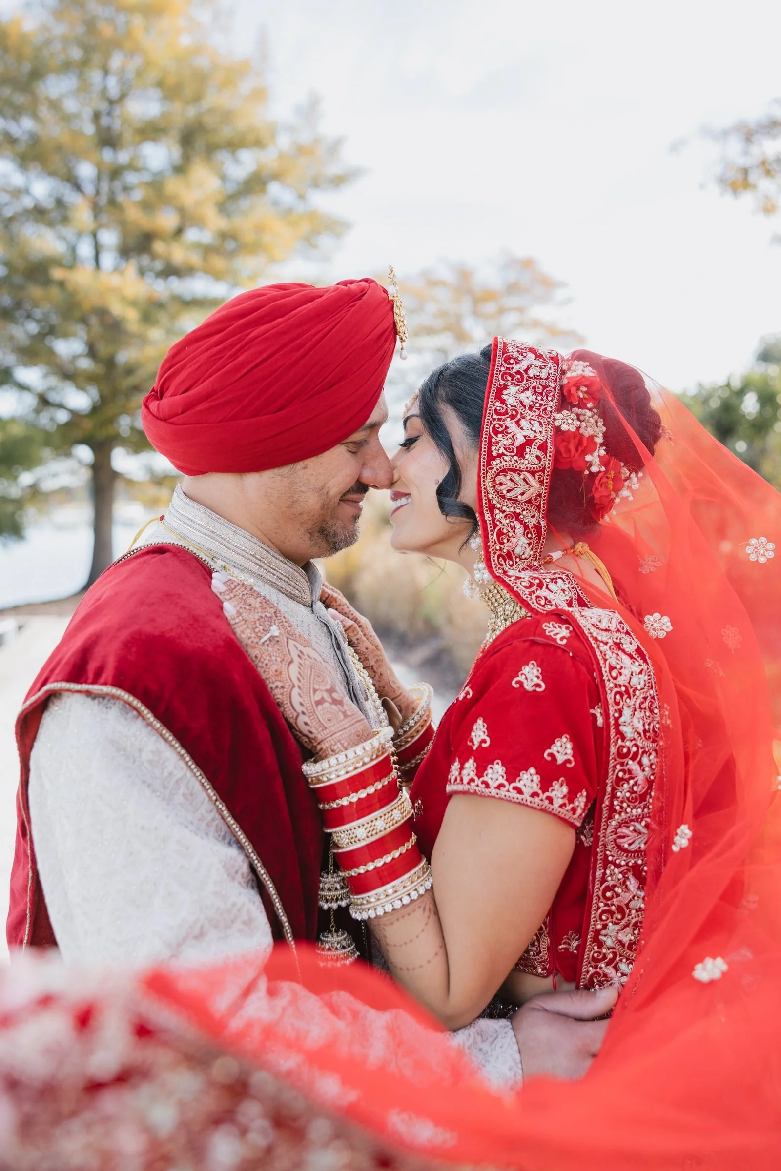 A couple dressed in traditional Indian wedding attire face each other closely, smiling with their foreheads touching. The man is wearing a red turban and cream-colored kurta with intricate embroidery, while the woman is dressed in a red saree with go