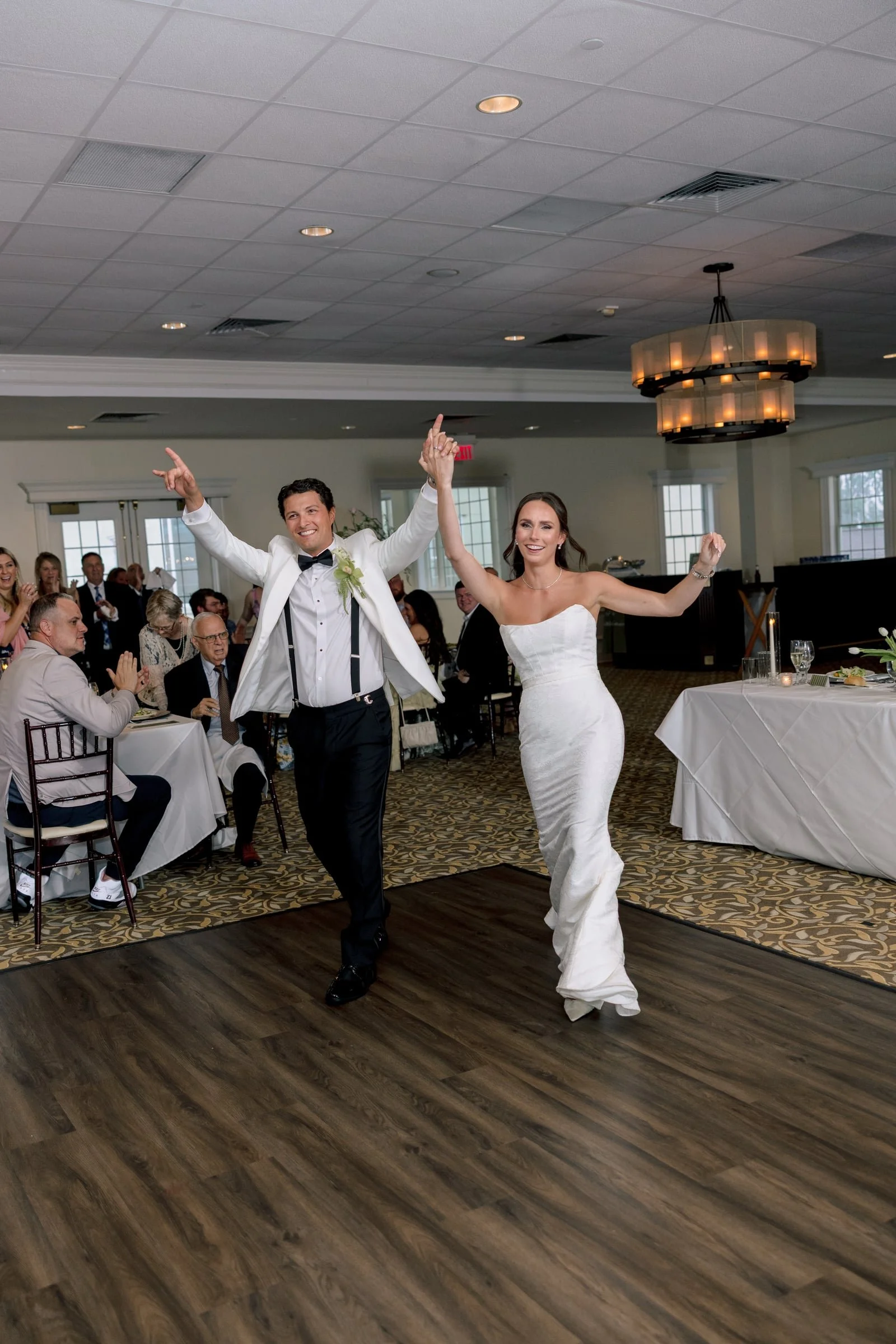A bride and groom dancing together at their wedding reception, holding hands and smiling. The bride is wearing a strapless white wedding gown, and the groom is in a white tuxedo jacket with a black bow tie. Guests are sitting at tables in the backgro