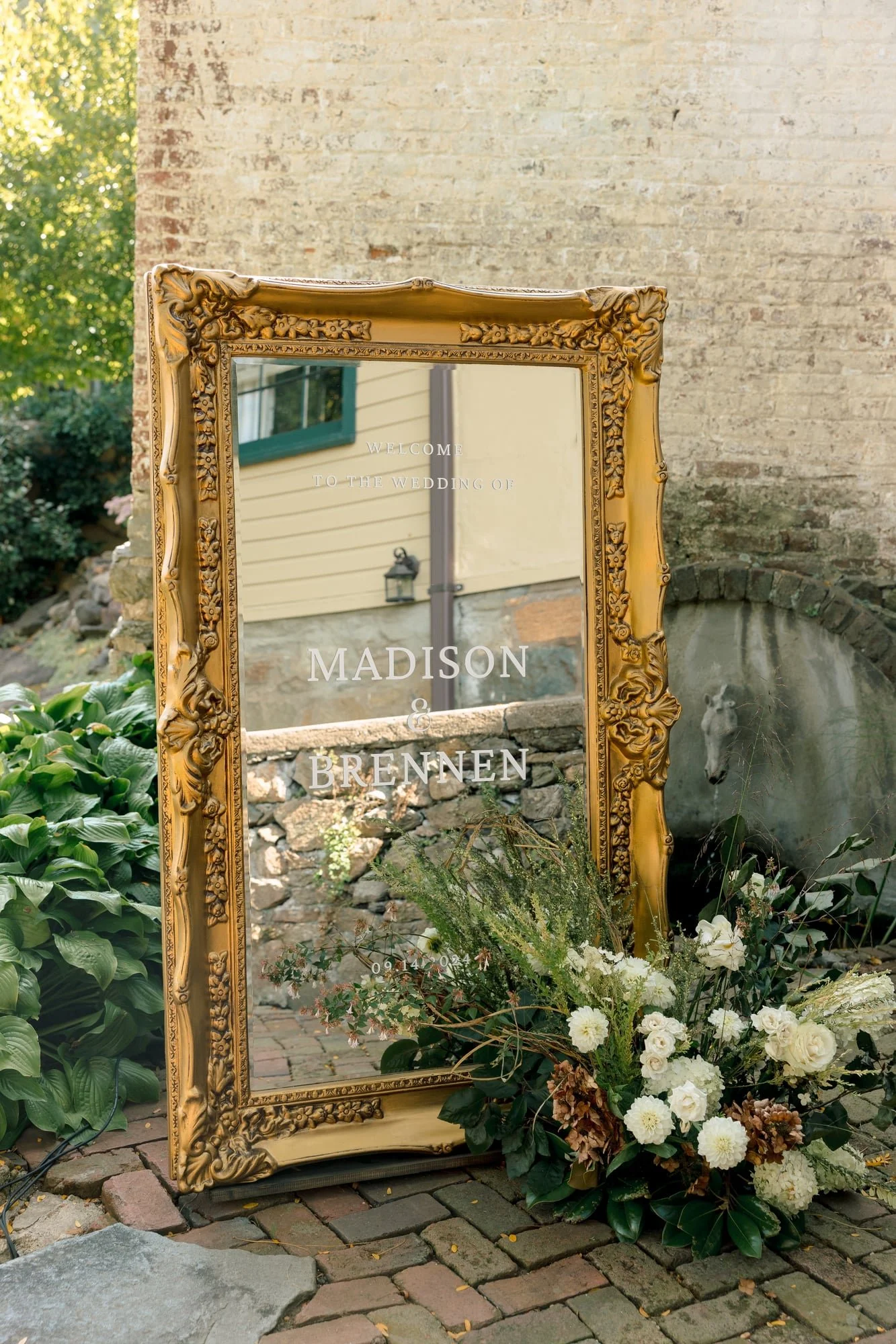 Decorative gold-framed mirror with wedding welcome text, surrounded by white flowers and greenery, outdoors with brick paving and a brick building in the background.