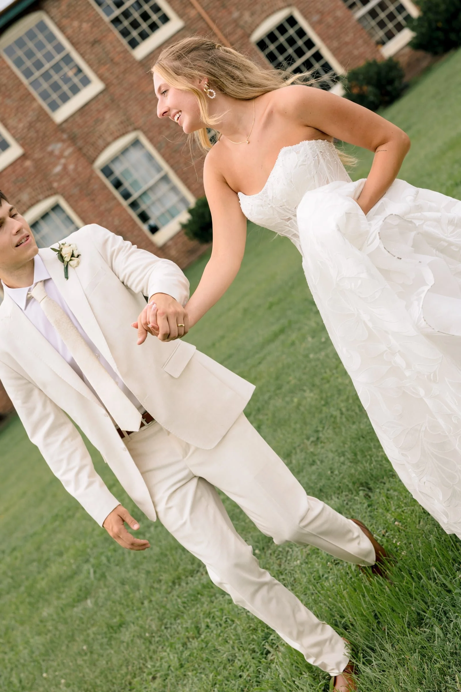 A bride in a strapless white wedding gown holding hands with a groom in a cream-colored suit outdoors on a grassy area with a brick building in the background.