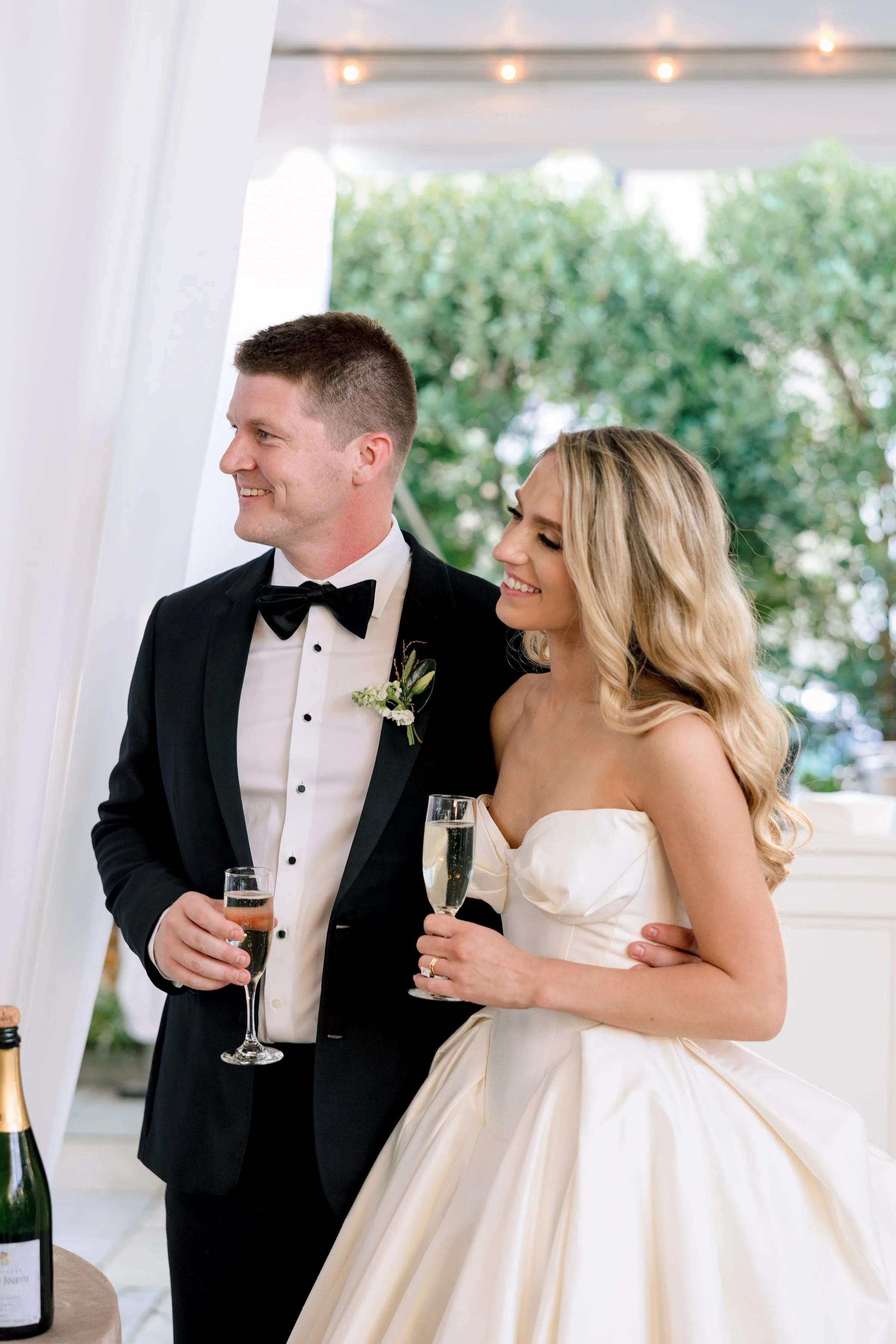 A newlywed couple dressed in wedding attire celebrating with champagne at their wedding reception. The groom wears a black tuxedo with a bow tie and boutonniere, and the bride wears a strapless white gown with her hair styled in loose waves. They are