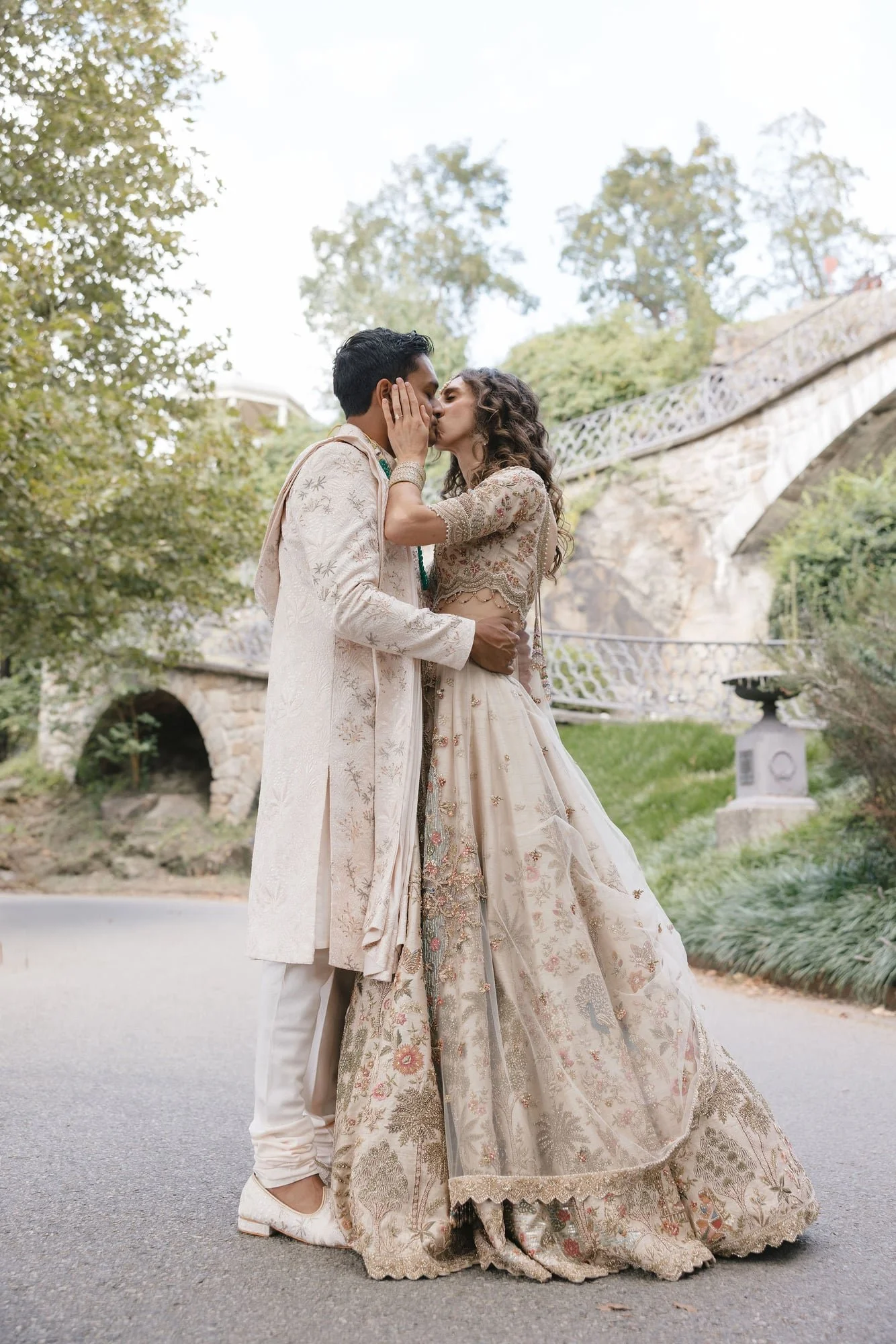 A man and woman dressed in traditional Indian wedding attire kiss in an outdoor setting with trees and a stone bridge in the background.