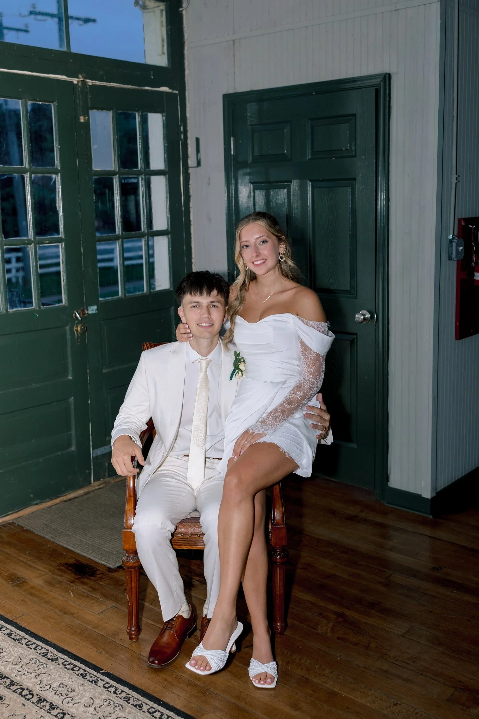 A young couple at their wedding, with the bride sitting on the groom's lap. The groom is seated on a wooden chair wearing a white suit and cream tie, while the bride is dressed in a white wedding gown with off-the-shoulder lace sleeves. They are indo