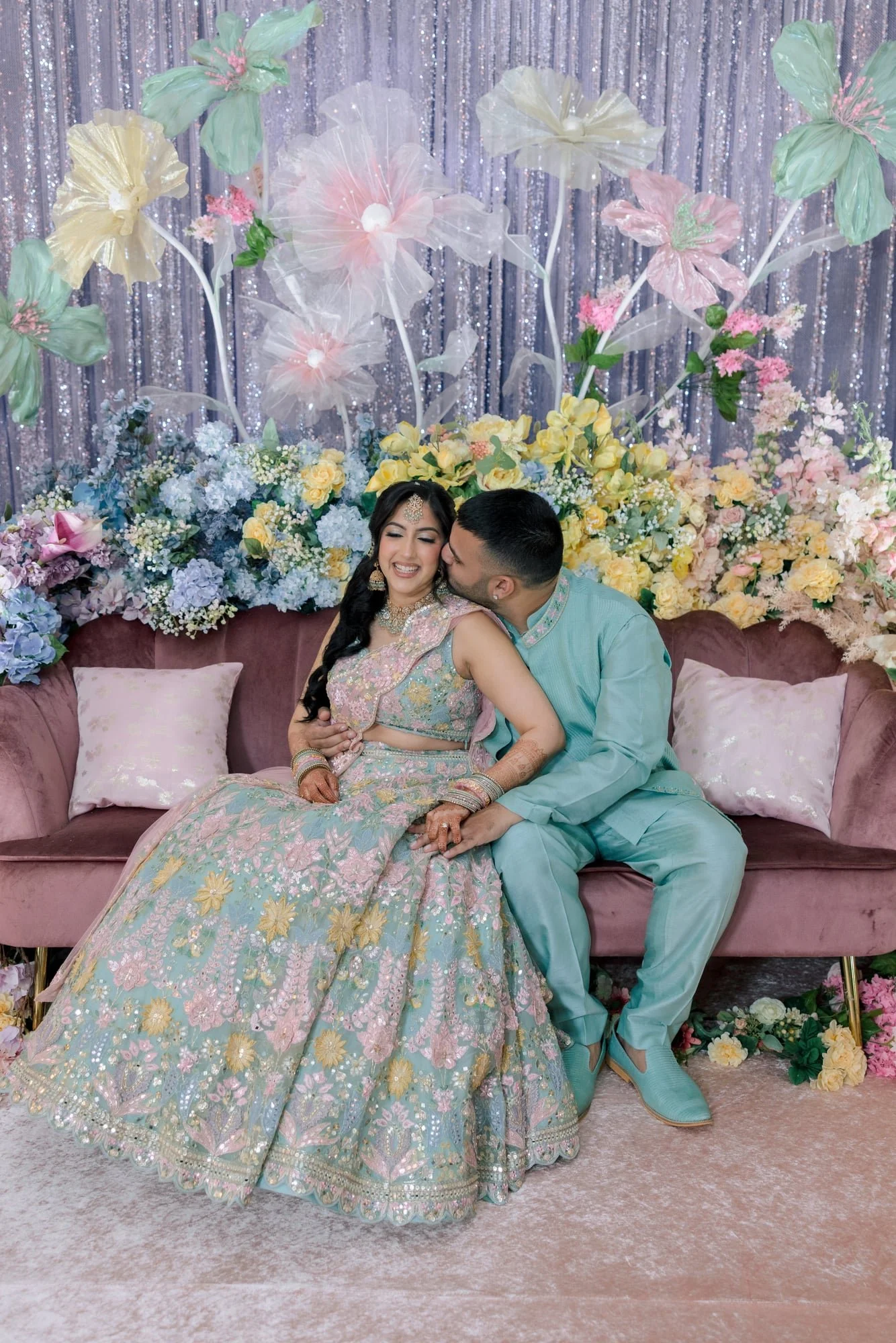 A bride and groom sitting on a pink velvet couch during their wedding celebration. The bride is wearing a pastel embroidered dress with floral patterns and traditional jewelry, while the groom is in a light teal traditional suit and shoes. Behind the