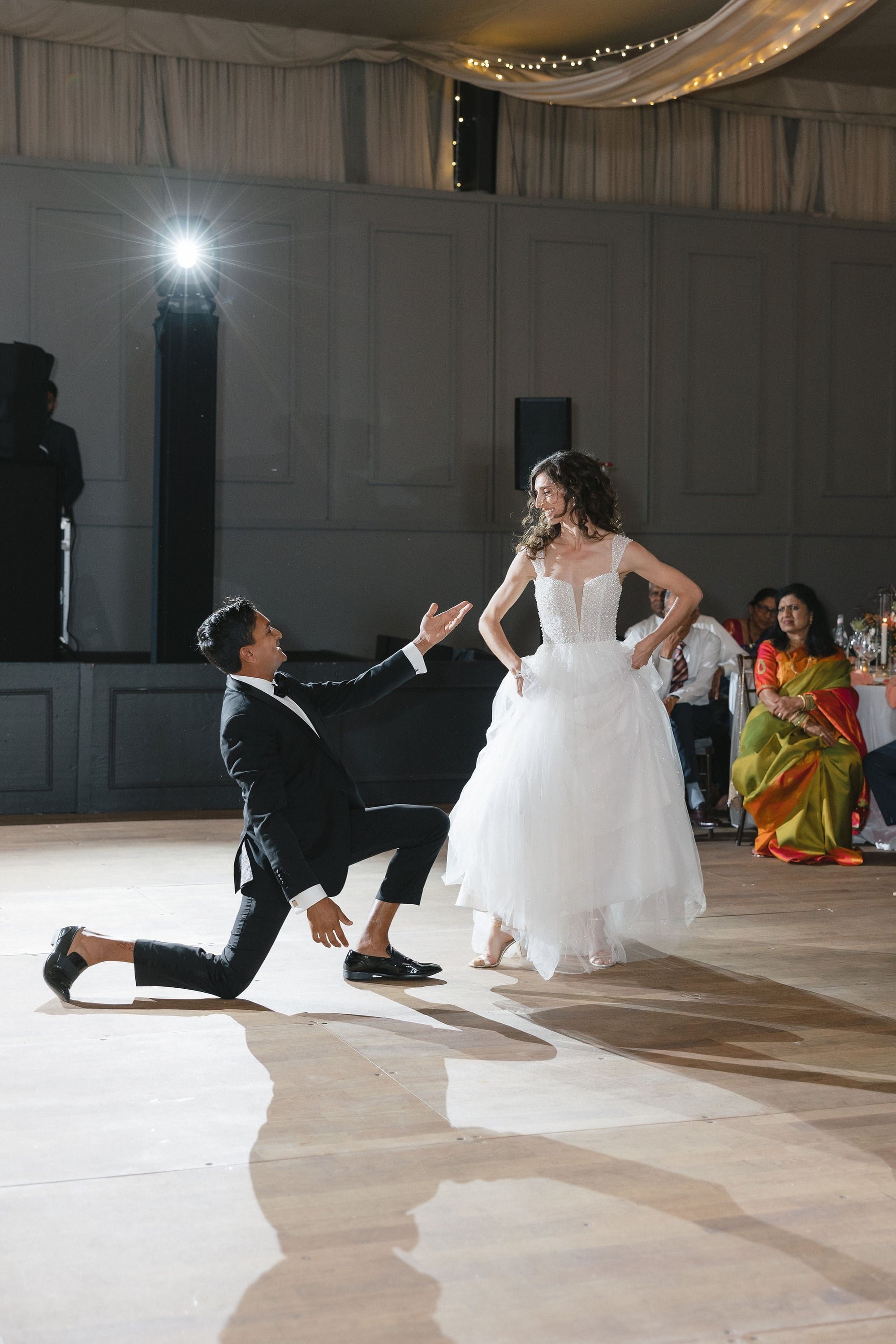 A man in a tuxedo kneels on one knee with a hand reaching up towards a woman in a wedding dress during a dance at a wedding reception. There are guests seated at tables in the background, some wearing colorful attire, in a decorated event hall with s