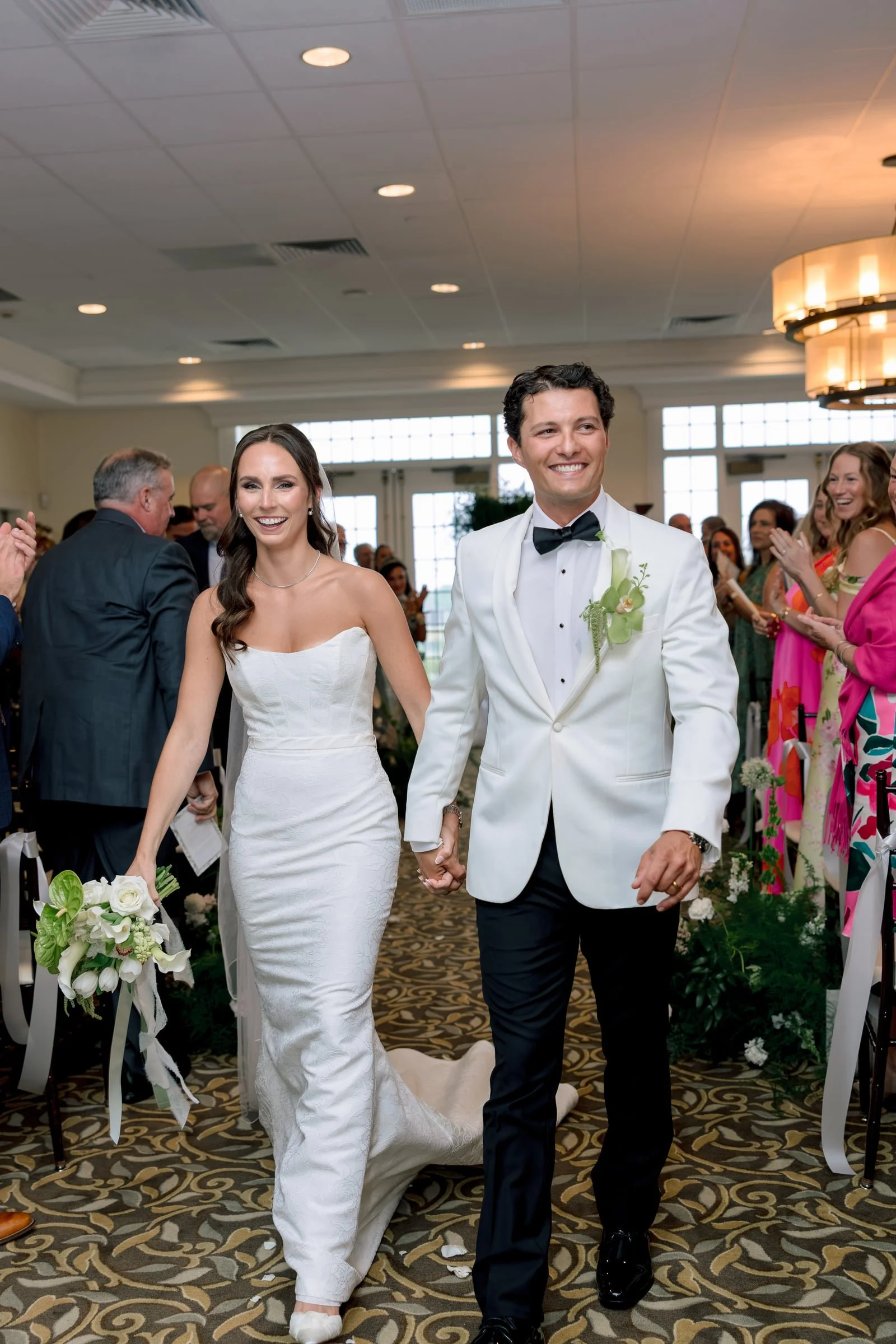 A bride and groom are walking down the aisle at their wedding reception, holding hands and smiling, with guests clapping and celebrating around them.