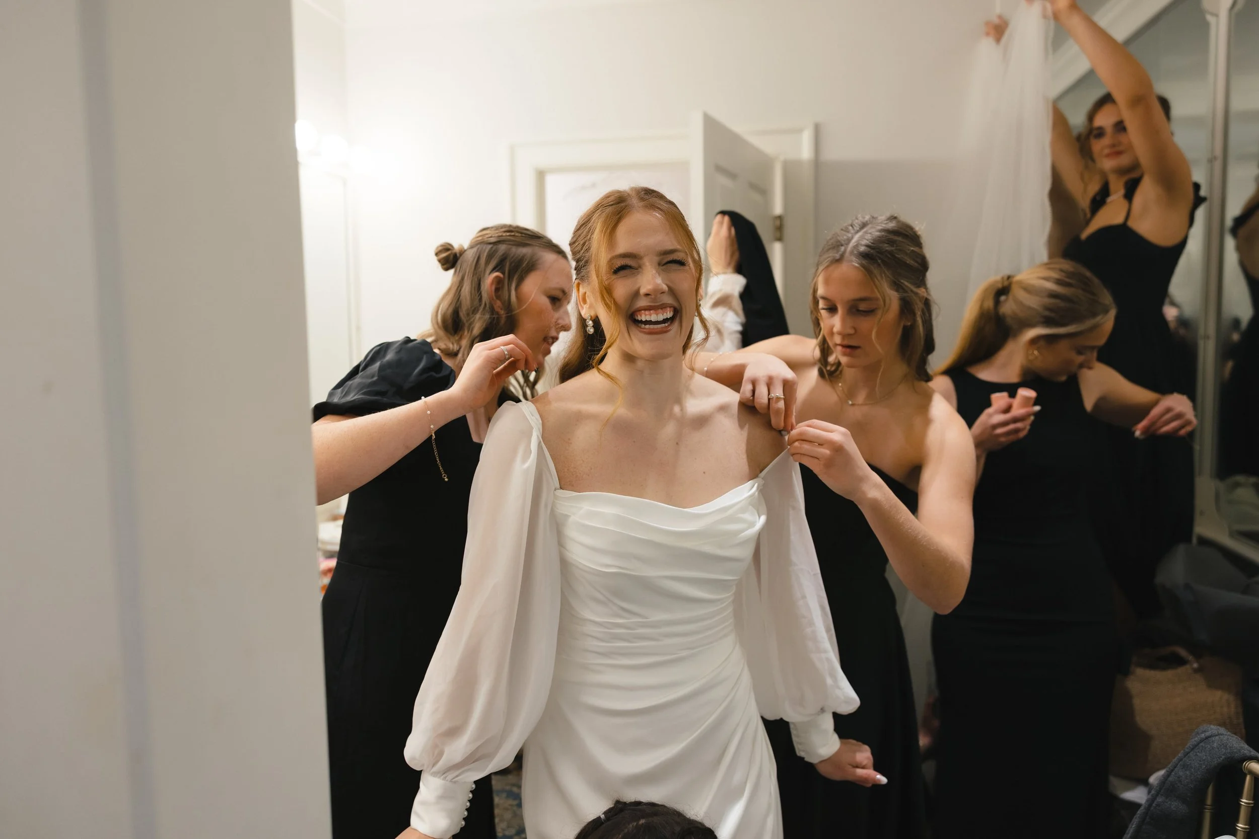 A woman wearing a white dress is smiling and getting ready, surrounded by friends in a dressing room, with one helping her with her dress and another adjusting her hair.