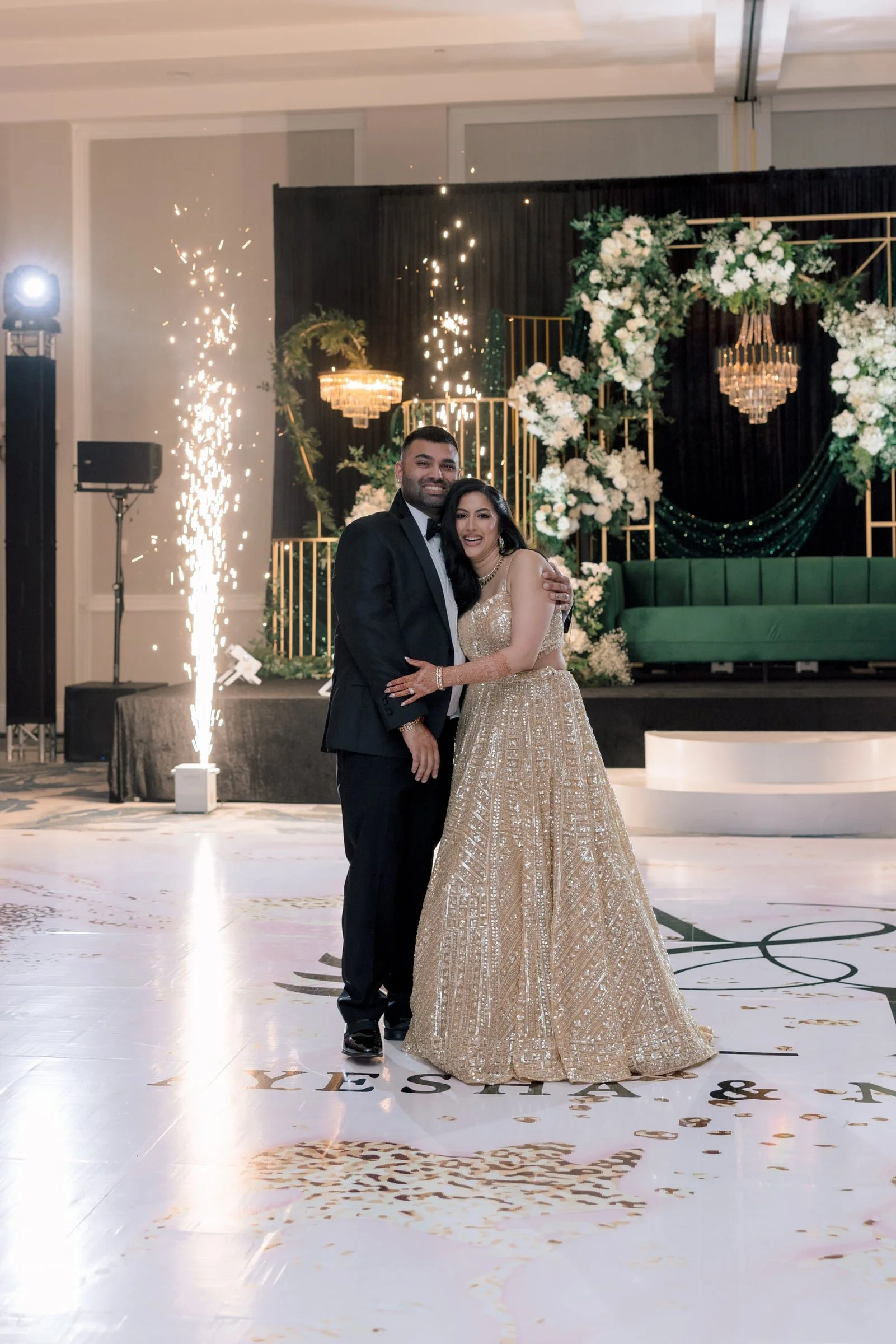 Couple in formal attire, the man in a tuxedo and the woman in a sparkling gold gown, standing on a wedding dance floor with a backdrop of floral decorations, chandeliers, and pyrotechnics.
