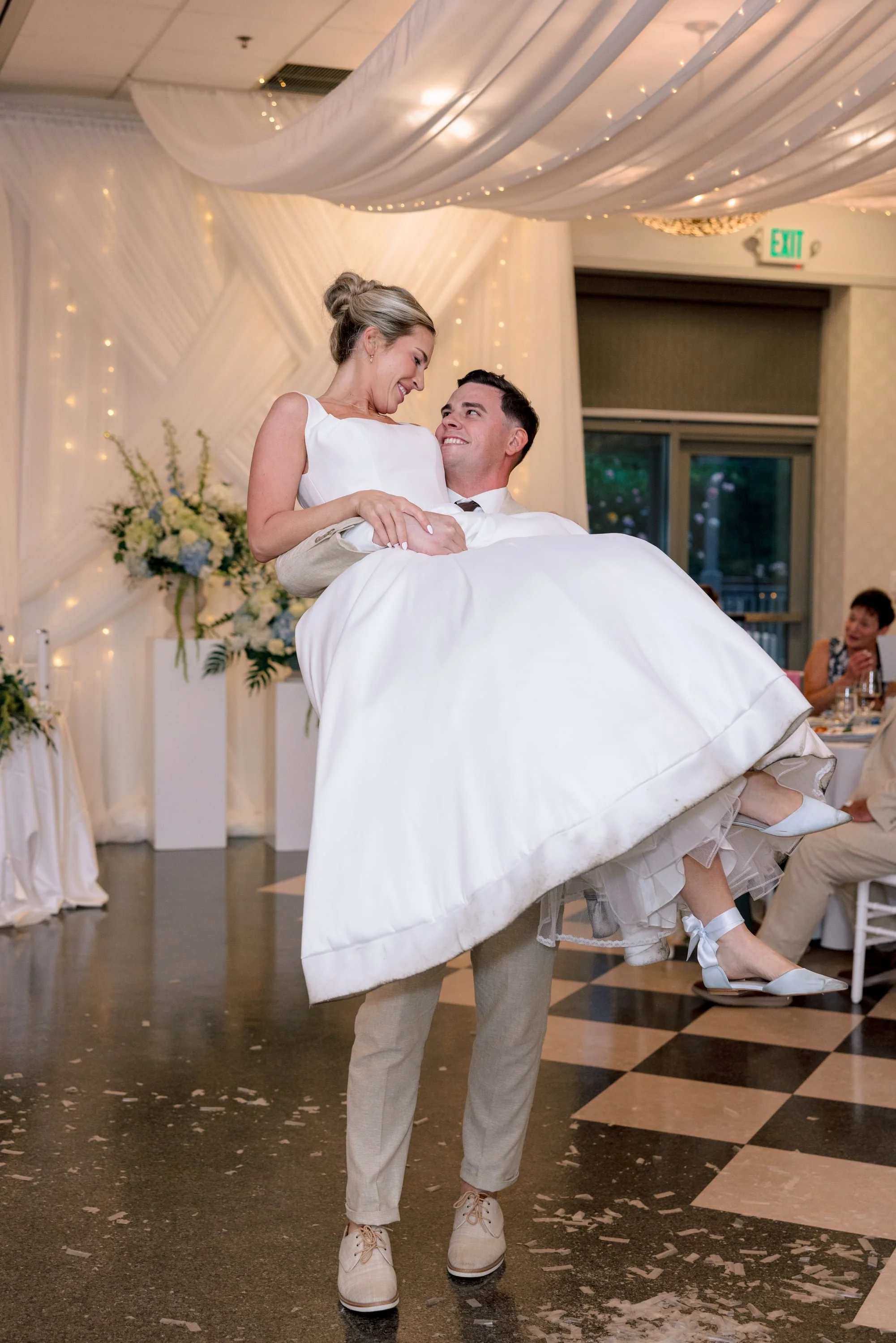 A man in a beige suit and white shirt is holding a woman in a white wedding dress and white shoes, dancing at a wedding reception with decorations, flowers, and guests in the background.