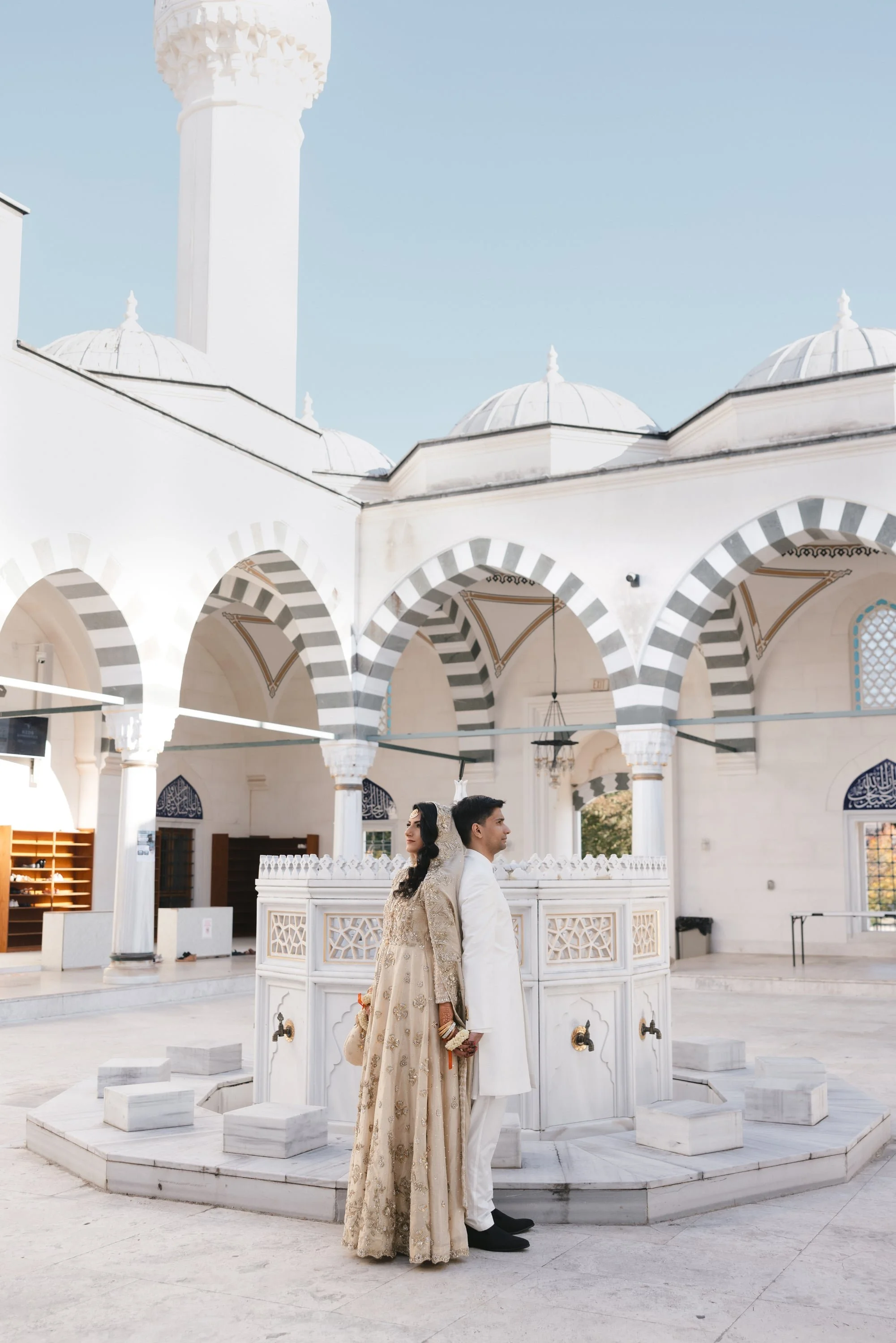 A bride and groom standing back to back in front of a white marble fountain inside a mosque with Islamic-style architecture and arches.