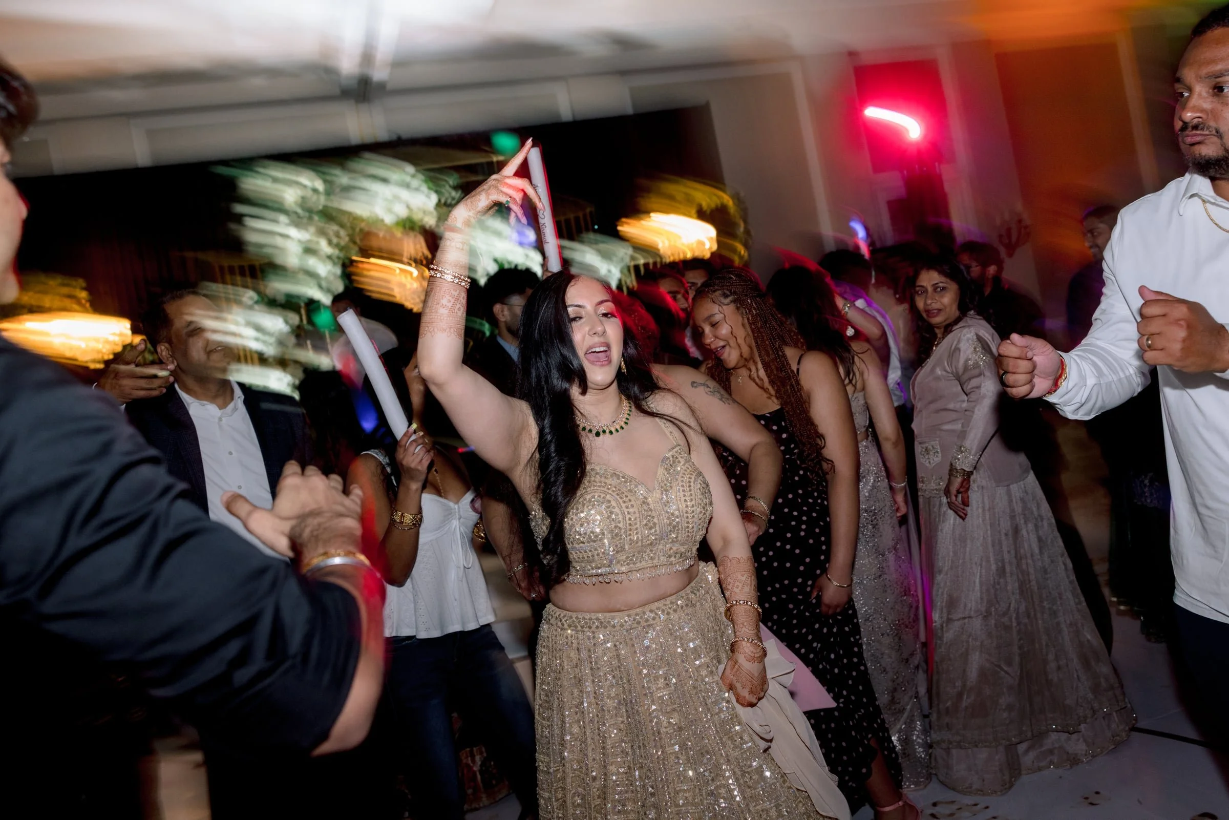 A group of people dancing at a celebration or party, including a woman in a gold traditional dress and henna on her hands, surrounded by others in festive attire, with colorful lights and motion blur.