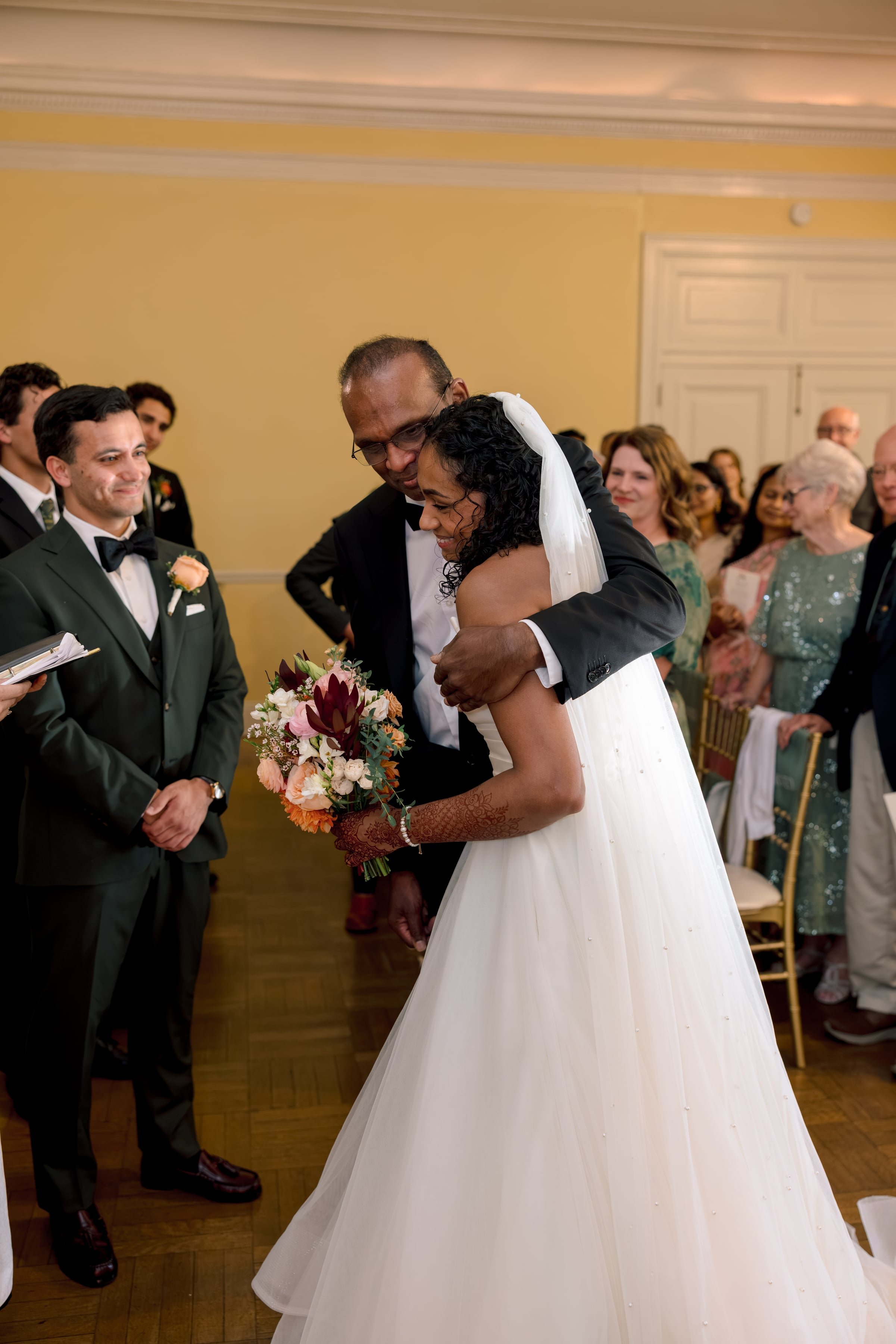 A bride and groom share a dance at their wedding reception. The bride is wearing a white gown and veil, holding a bouquet of flowers, while the groom is dressed in a black tuxedo. They are surrounded by guests watching and smiling.