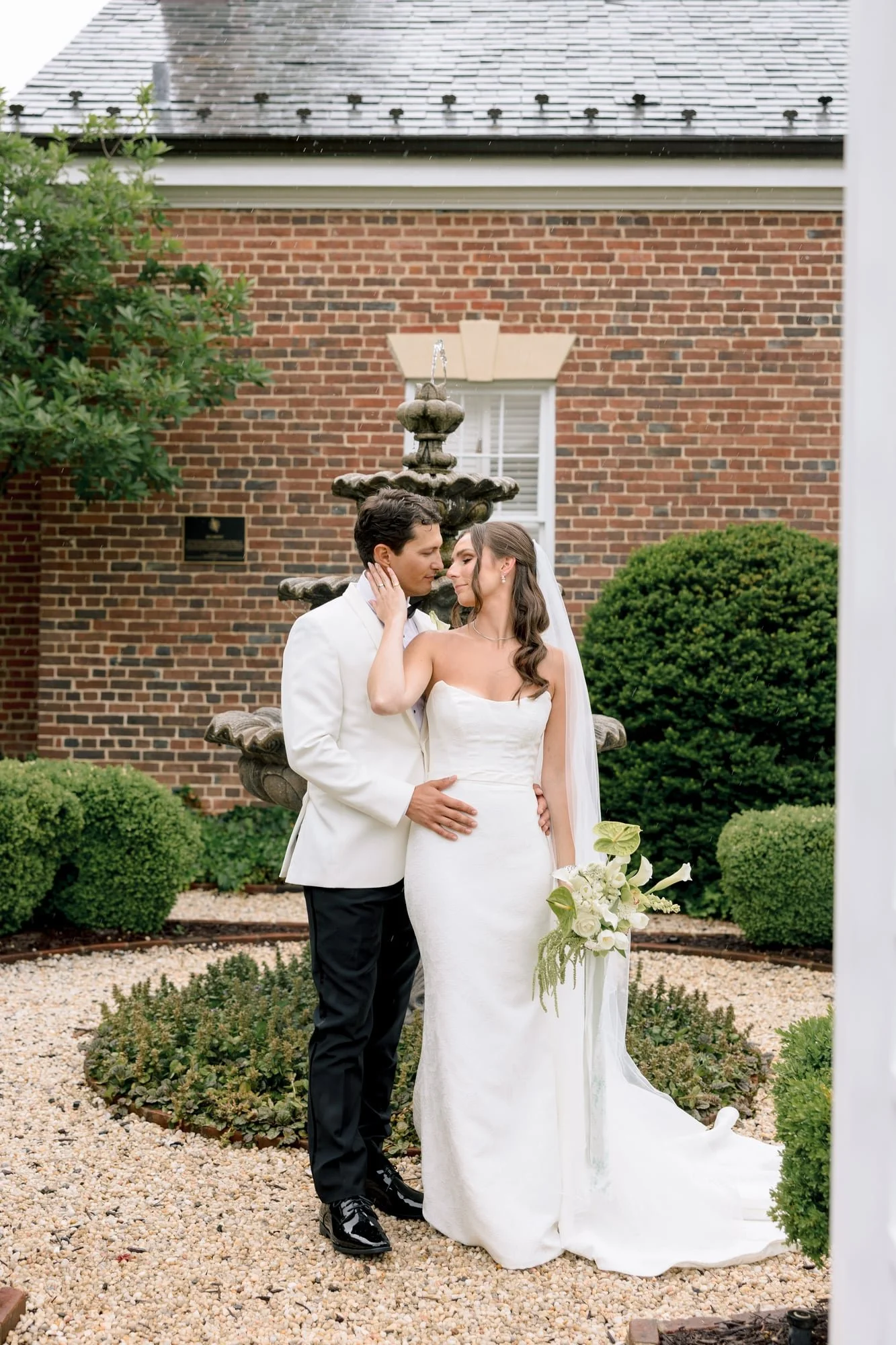 A newlywed couple standing close together outdoors in front of a brick building with a decorative fountain, greenery, and a window in the background. The groom is wearing a white tuxedo jacket and black pants, and the bride is in a strapless white wedding gown holding a bouquet of white flowers. They are looking into each other's eyes affectionately.