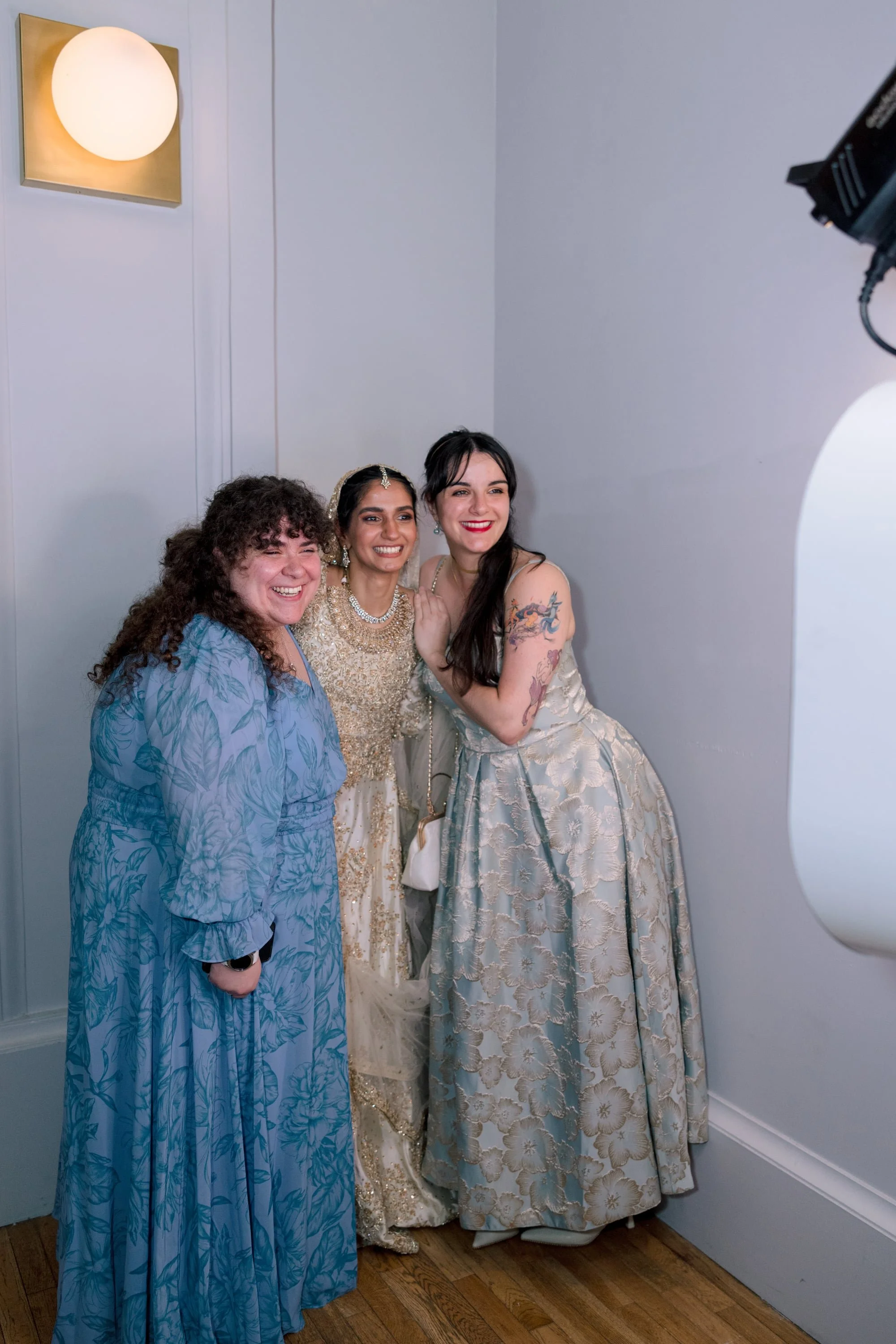 Three women smiling for a photo in a room with gray walls and a wooden floor. One woman is wearing a blue floral dress, the second is in a gold embroidered gown with jewelry, and the third is in a cream-colored dress with floral patterns and tattoos 