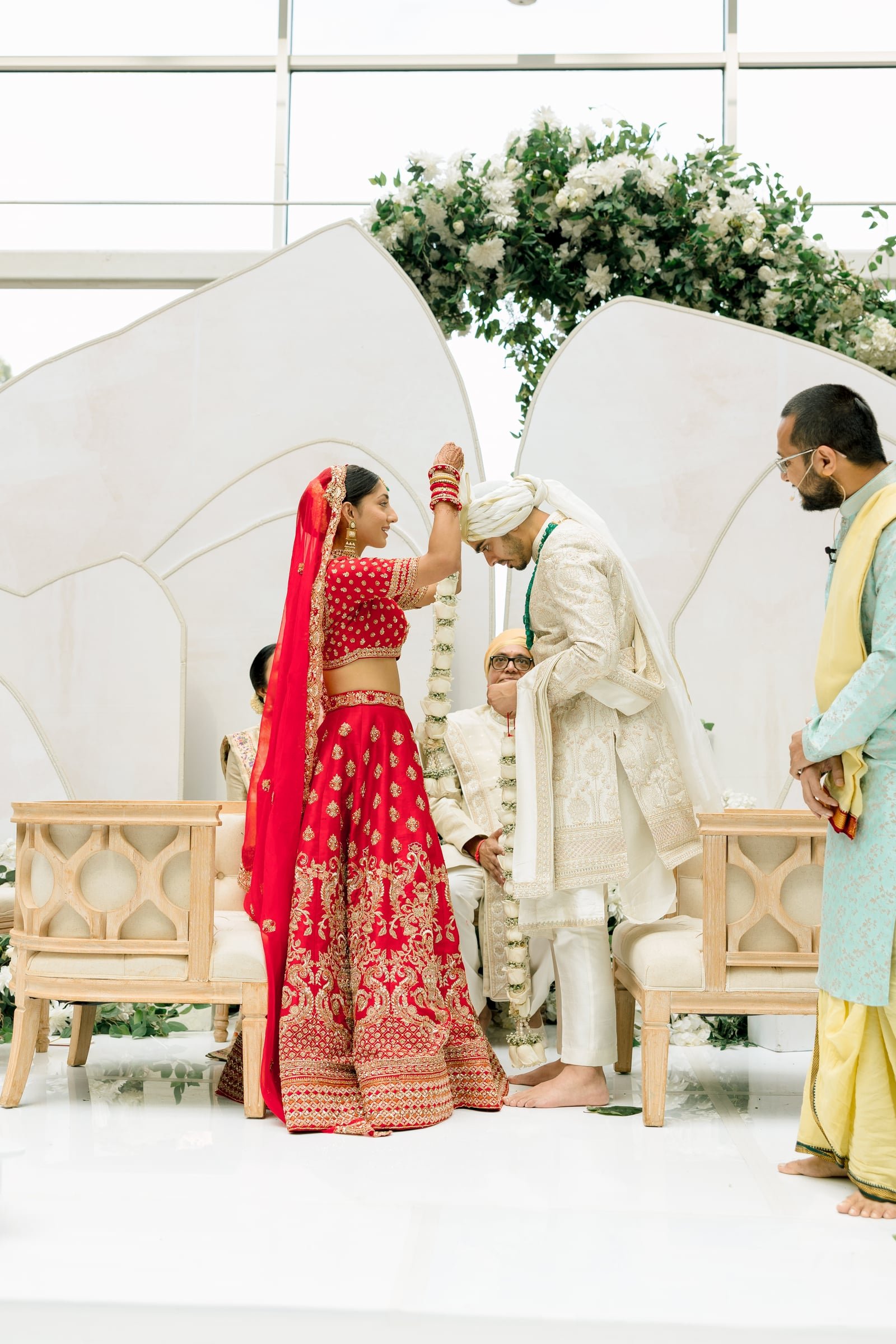A traditional Indian wedding ceremony with the bride and groom dressed in ornate attire, exchanging vows under a floral arch, with family members present.