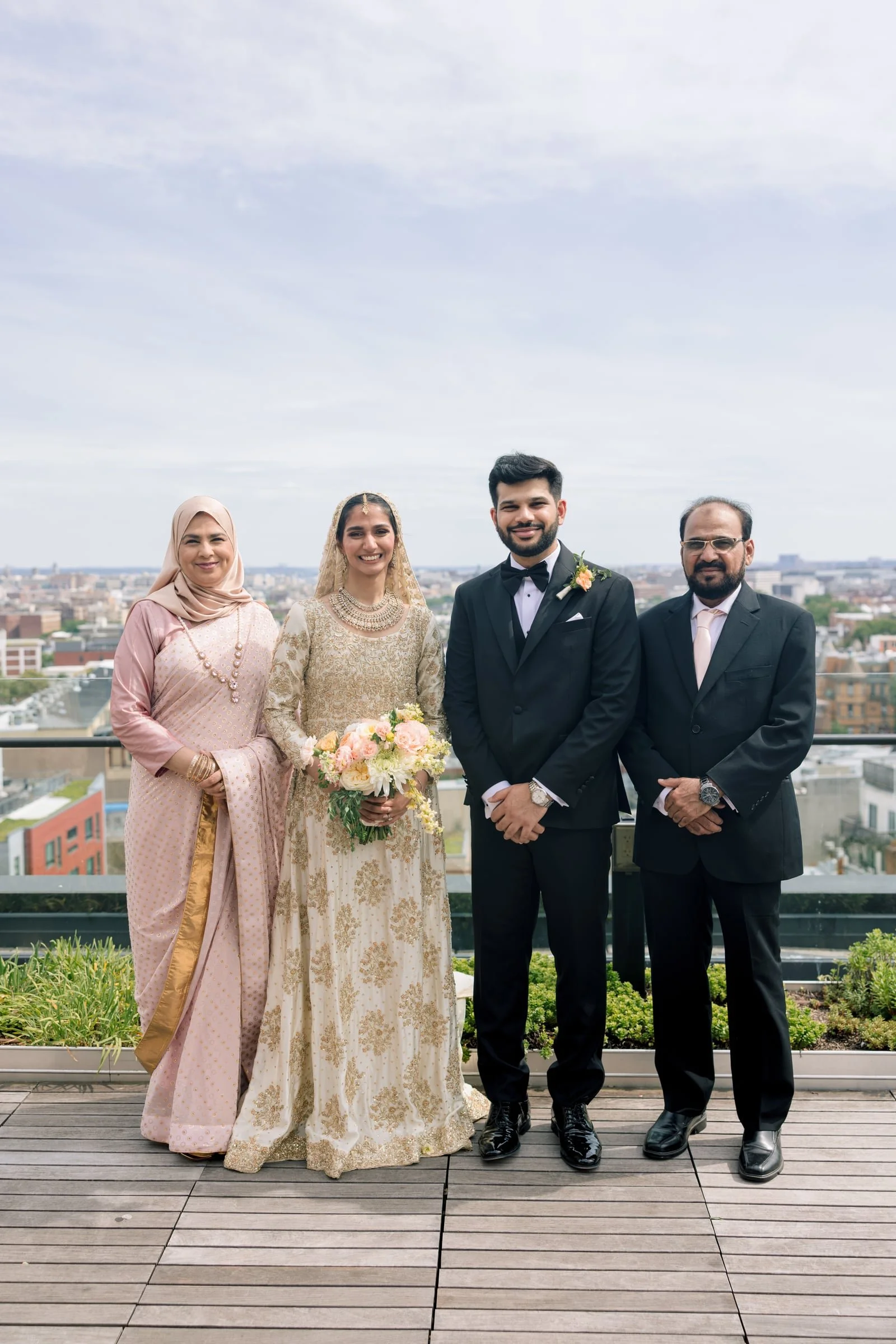 Four people standing together on a rooftop with a cityscape in the background. A woman in traditional South Asian attire holding a bouquet of flowers, two men in suits, and a woman in a traditional outfit. All are smiling.