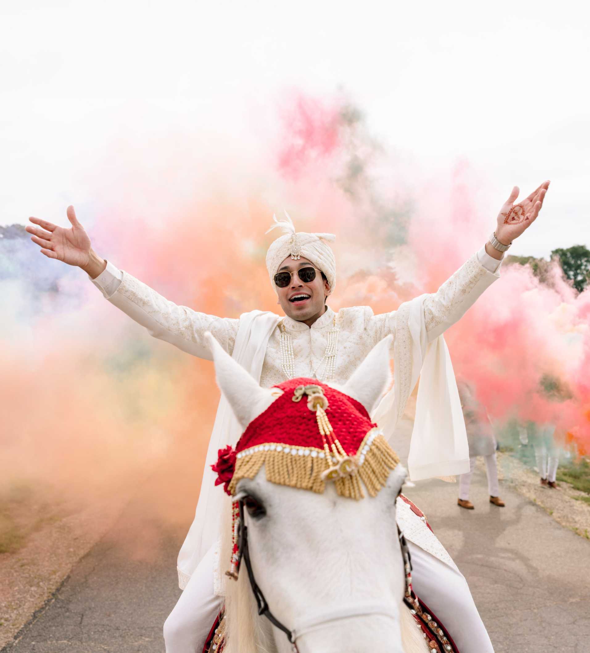 Person wearing traditional Indian attire riding a white horse, with arms raised, colorful powder in the background, during a celebration or festival.