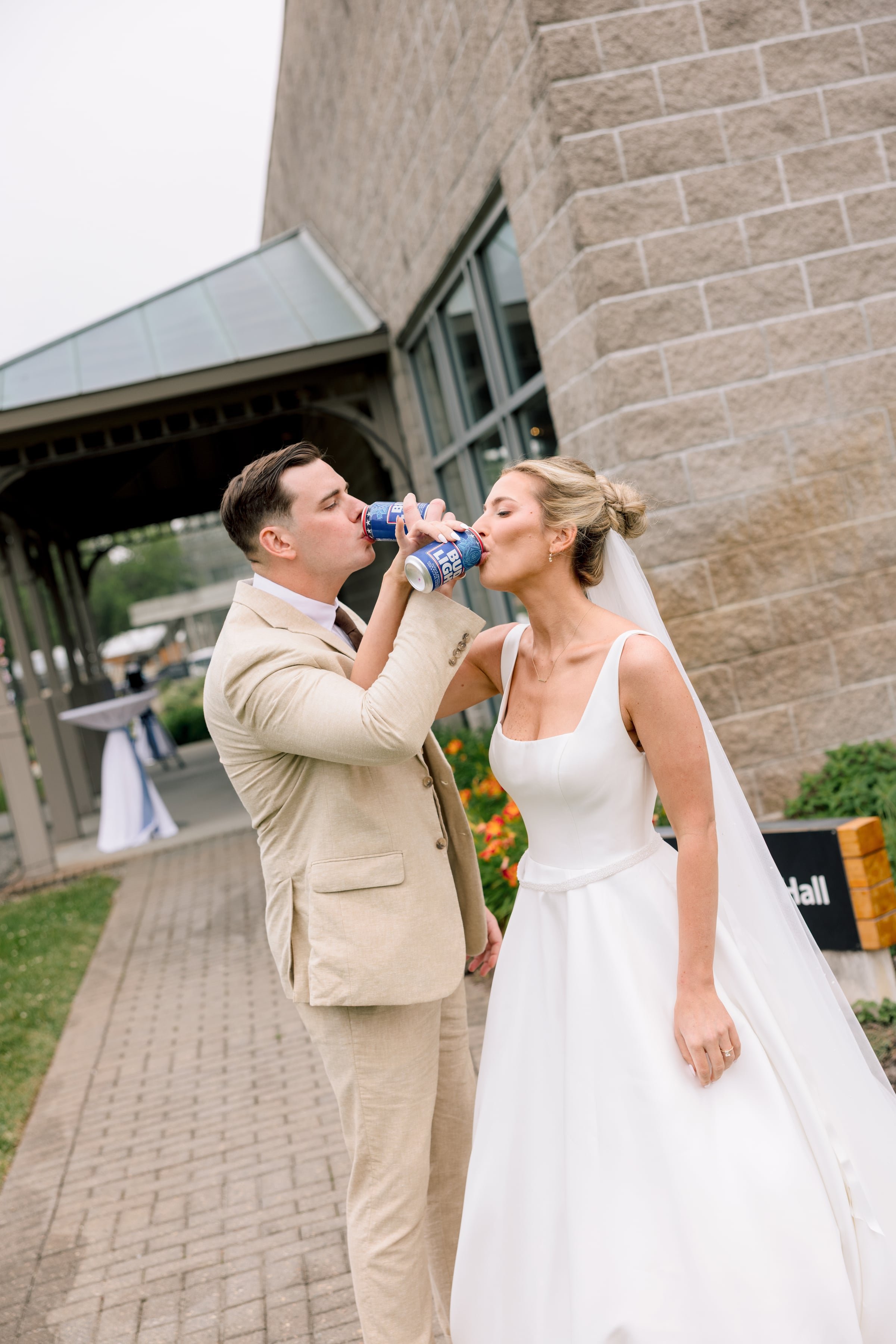 A newlywed couple in wedding attire sharing drinks at an outdoor event, standing on a brick walkway outside a building with large windows and brick walls.