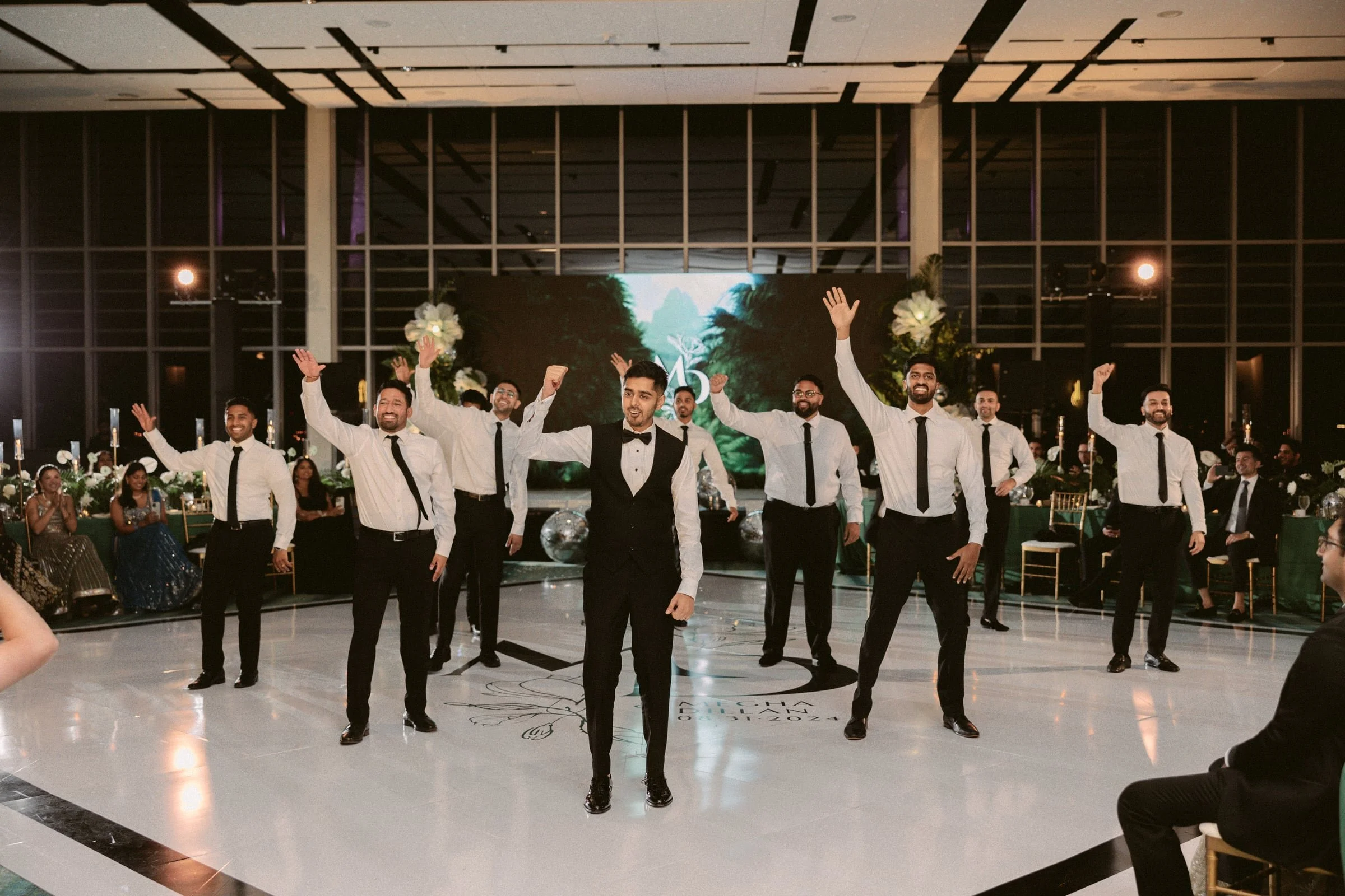 A group of seven men dressed in tuxedos and white shirts performing a dance on a ballroom floor with a decorated background and seated guests watching.