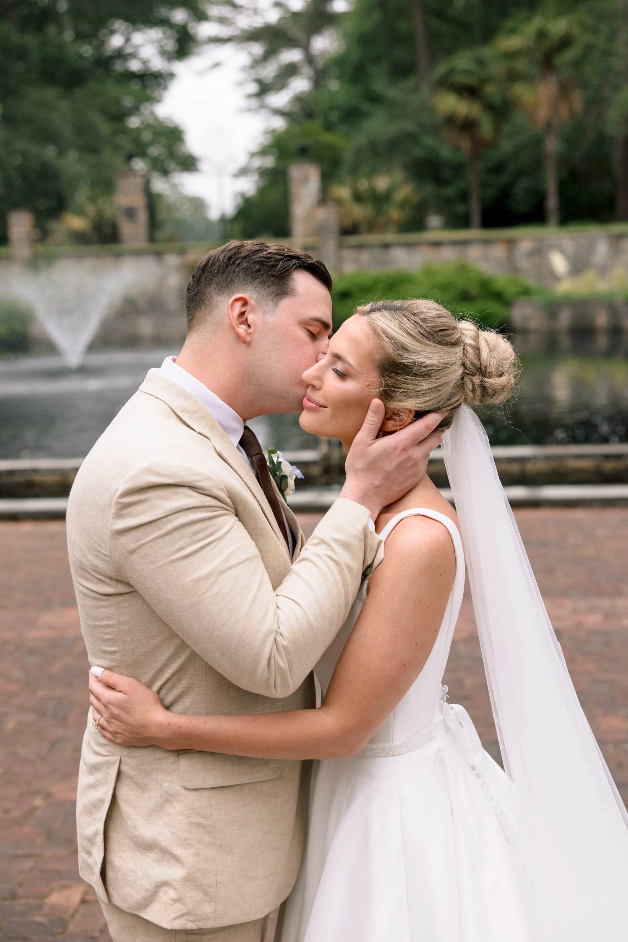 A newlywed couple sharing a kiss outdoors near a fountain and greenery.