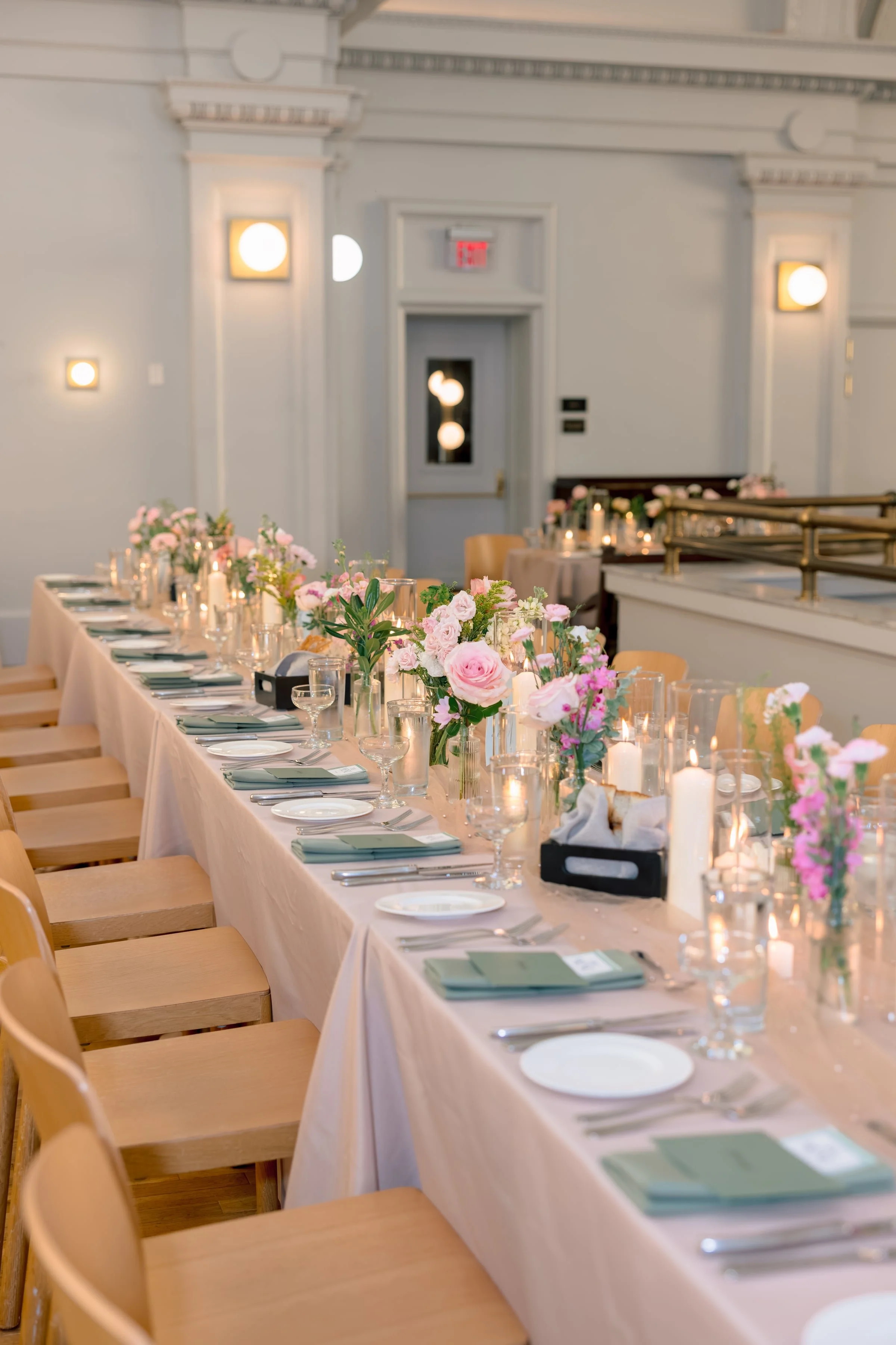 A long banquet table decorated with pink and white flowers, candles, and elegant tableware, set up for a formal event in a bright, white-walled room with wall-mounted lights and an elevated platform.