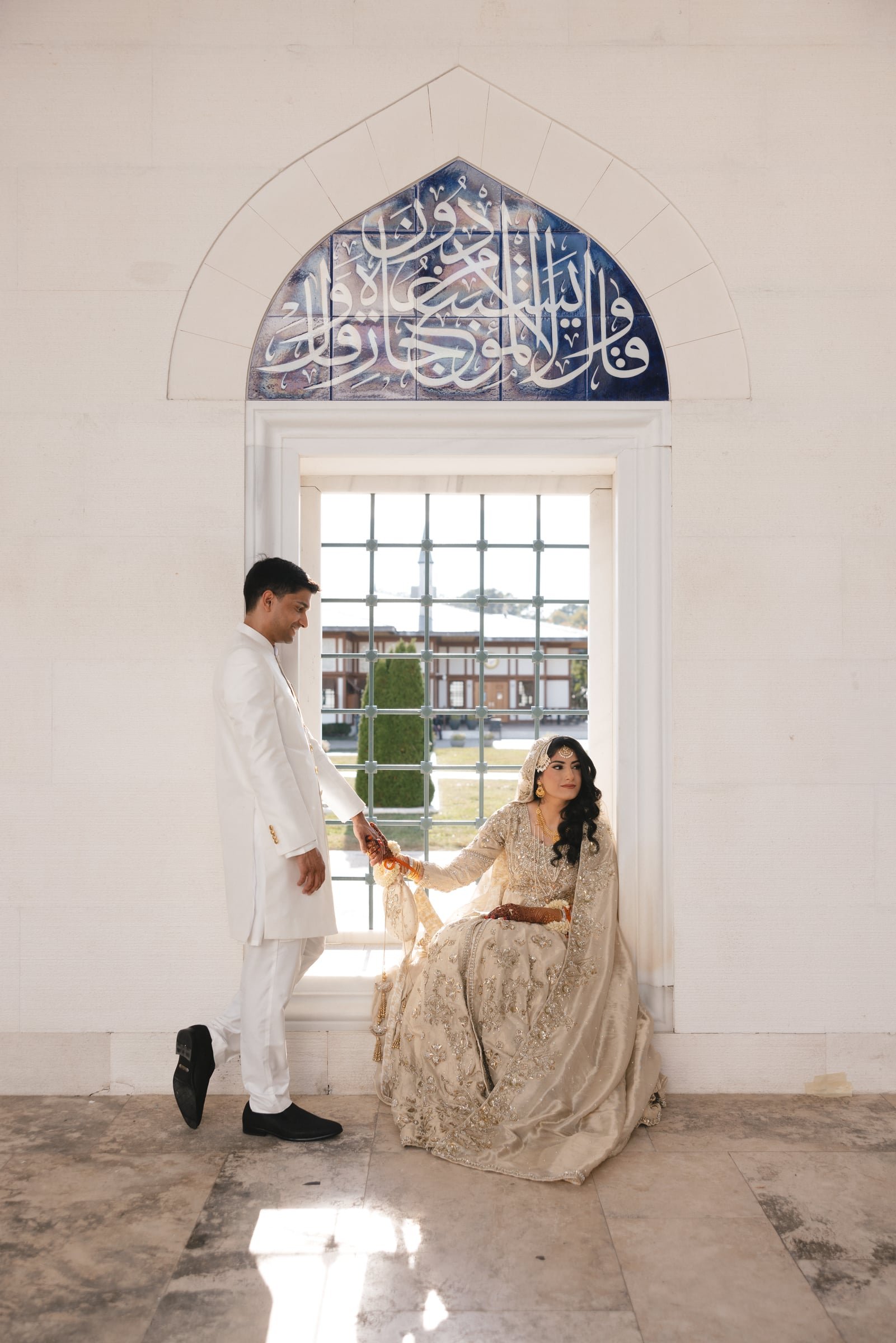 A bride dressed in an ornate gold and cream traditional wedding gown seated by a large window, holding a clutch, with a groom in a white suit standing beside her, holding her hand inside a bright, minimalistic room.