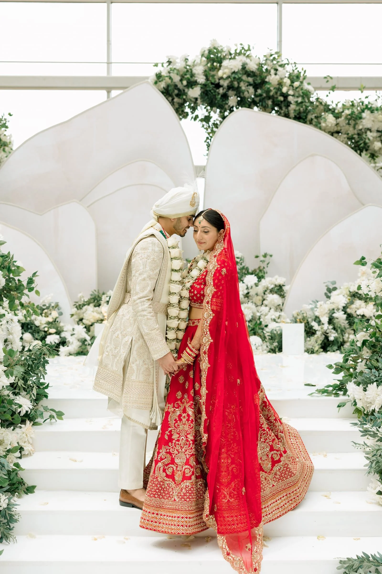 A bride and groom dressed in traditional Indian wedding attire, holding hands and leaning their foreheads together on a white staircase decorated with greenery and white flowers, with a floral arch in the background.