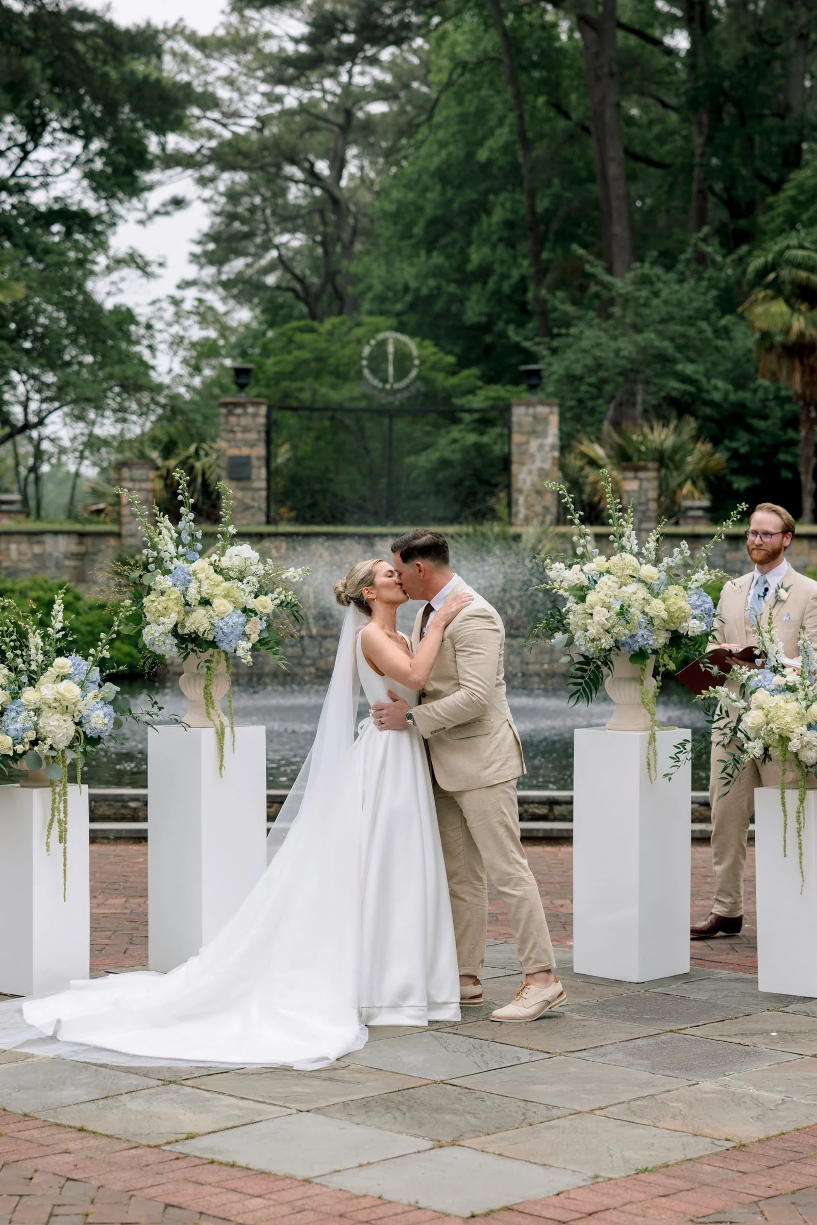A couple shares a kiss during their wedding ceremony in an outdoor garden setting with lush green trees, decorative floral arrangements, and a fountain in the background. The bride is wearing a white wedding dress with a long train, and the groom is dressed in a beige suit.