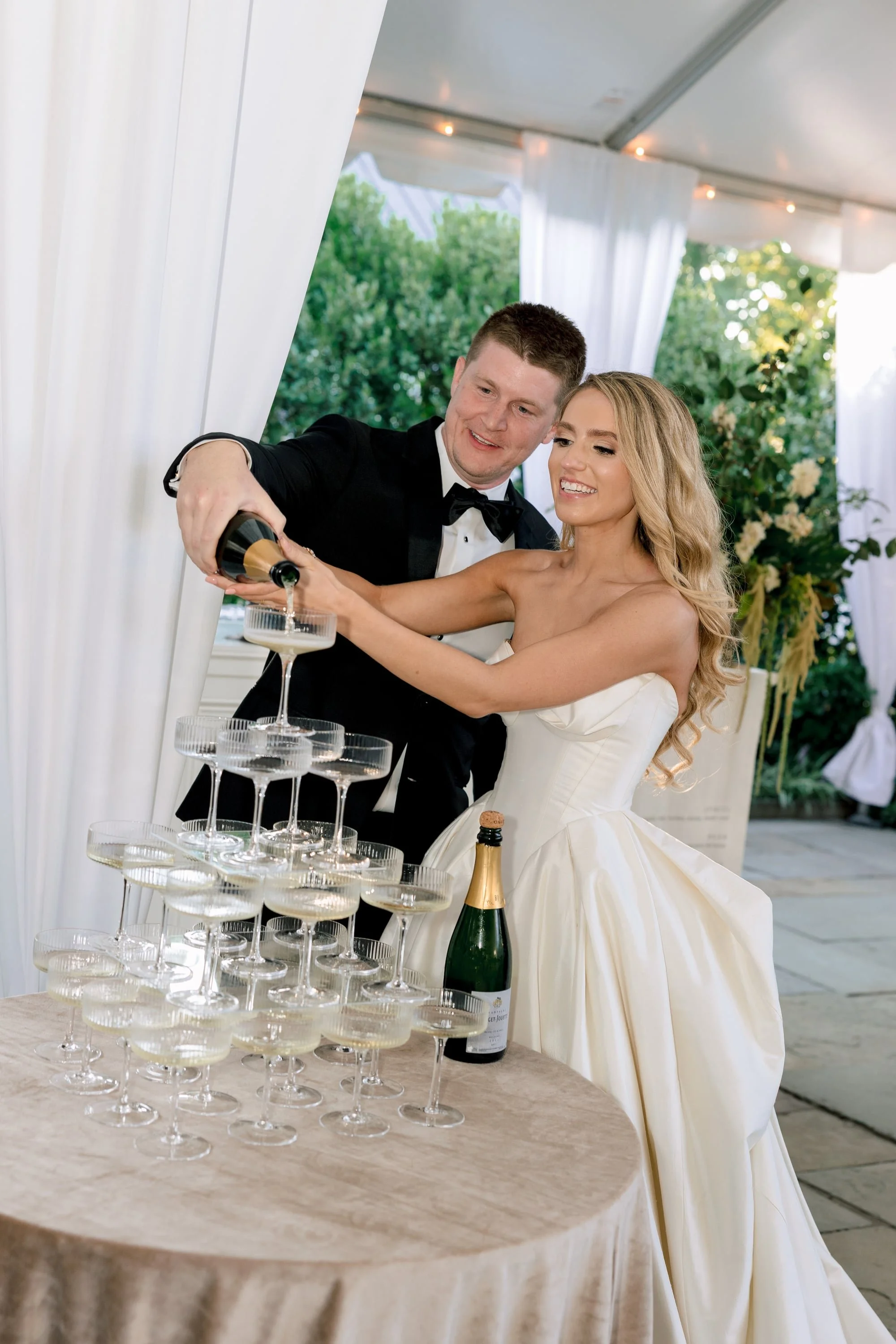 A bride and groom in wedding attire pouring champagne into a tower of coupe glasses at their wedding reception, decorated with white curtains and green foliage.