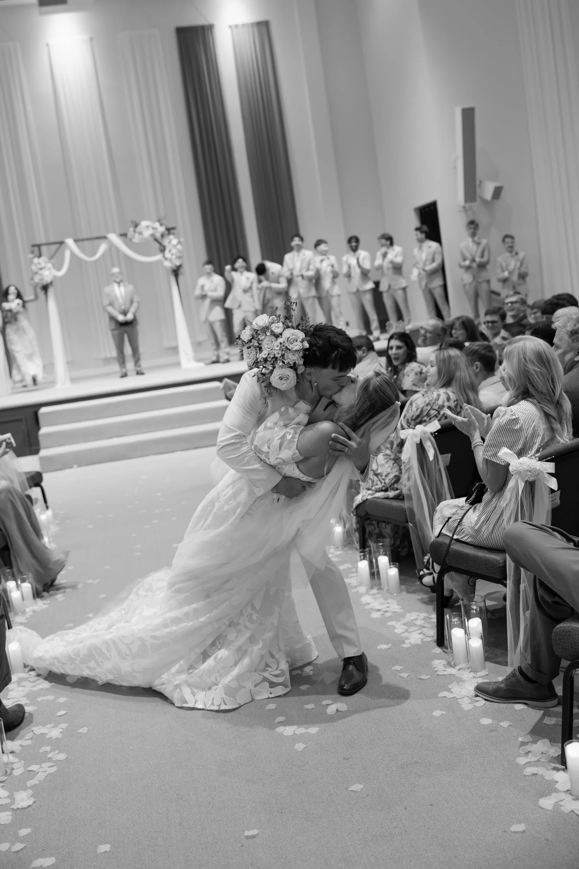 A bride and groom kiss in the aisle at their wedding ceremony with guests seated on either side and a decorated altar in the background.
