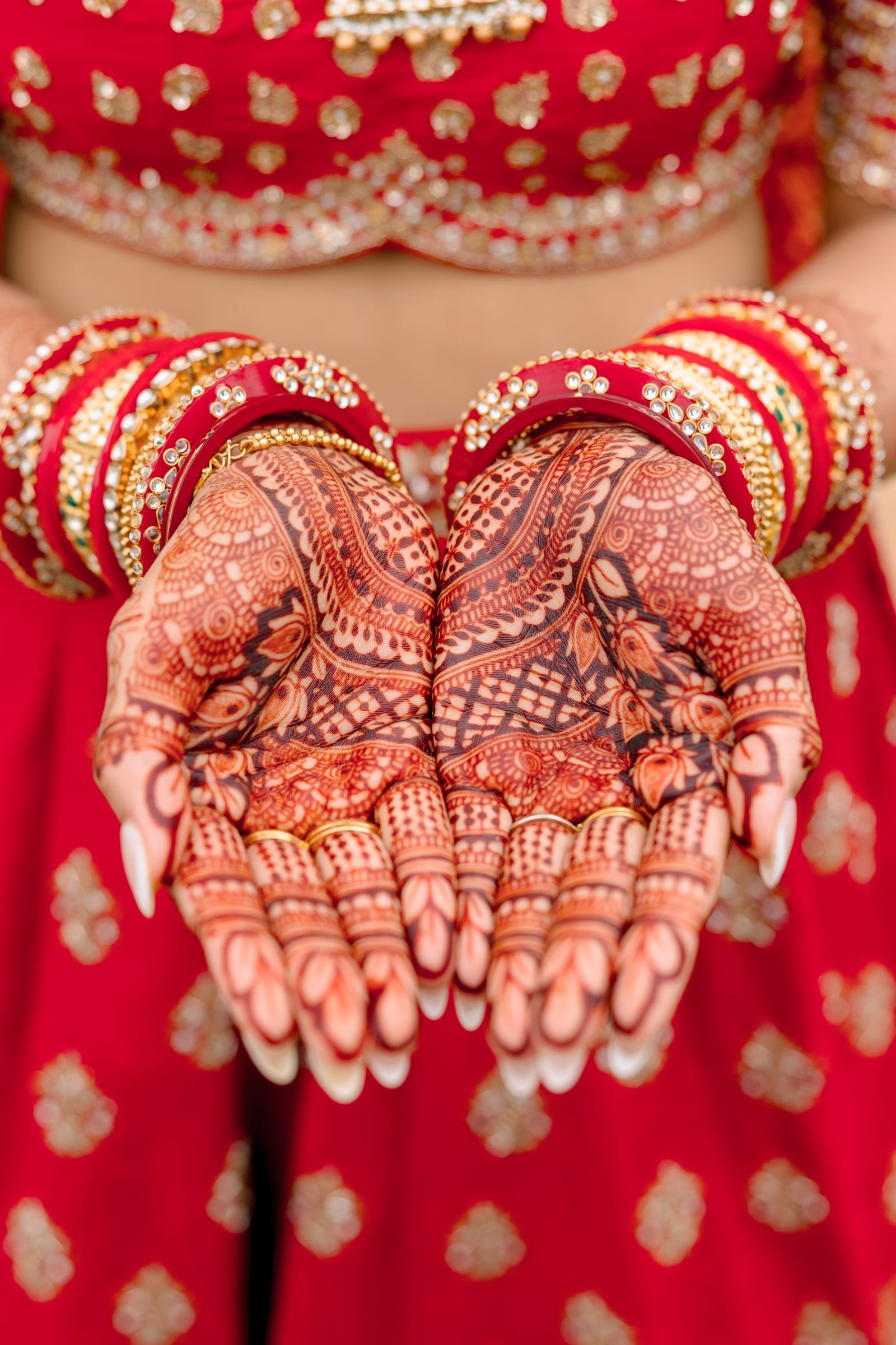 Close-up of hands holding a heart-shaped arrangement of red bangles decorated with rhinestones, with intricate henna designs on the hands, and a red outfit with gold embellishments in the background.