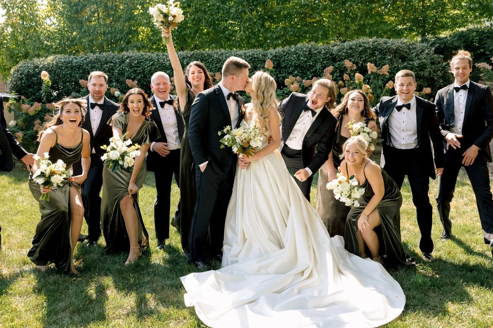 A wedding party outdoors with the bride and groom kissing in the center. The bride holds a bouquet and wears a white gown with a long train, while the groom wears a black tuxedo. Surrounding them are bridesmaids in matching dark dresses and groomsmen