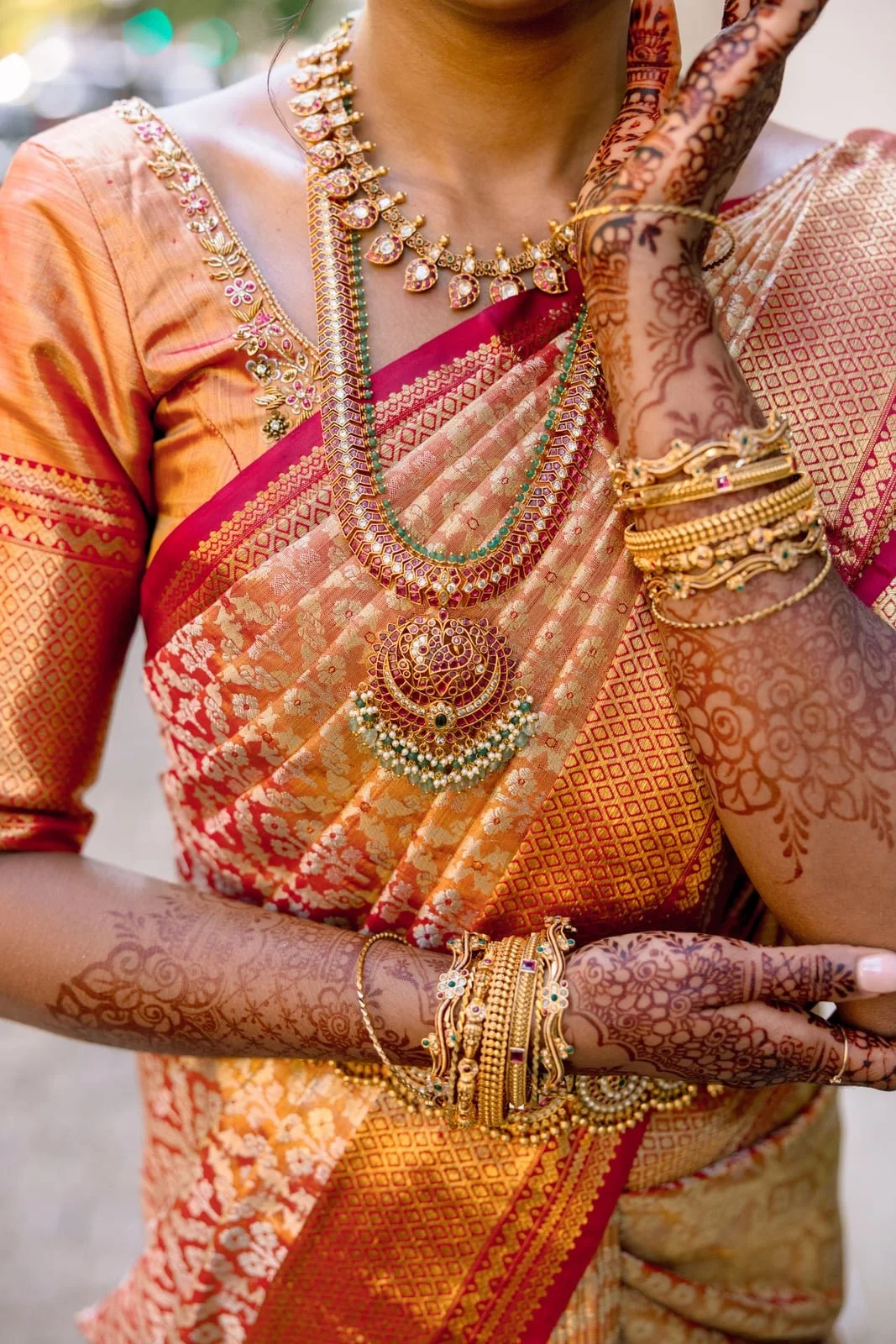 Close-up of a woman dressed in traditional Indian attire with gold jewelry, including necklaces, bangles, and rings, and elaborate mehndi (henna) designs on her arms and hands.