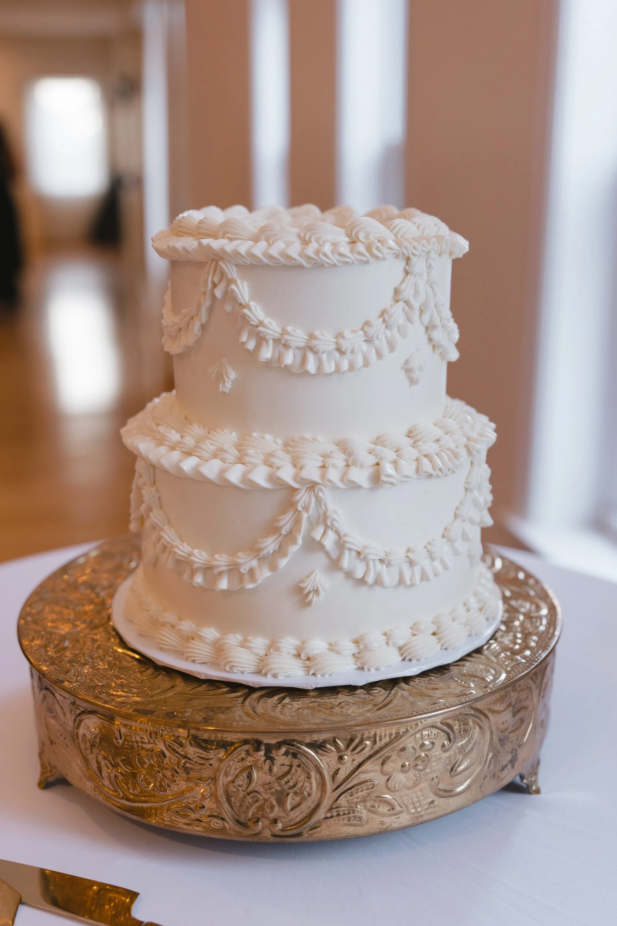 A three-tiered white wedding cake decorated with piped buttercream designs, sitting on an ornate gold cake stand.