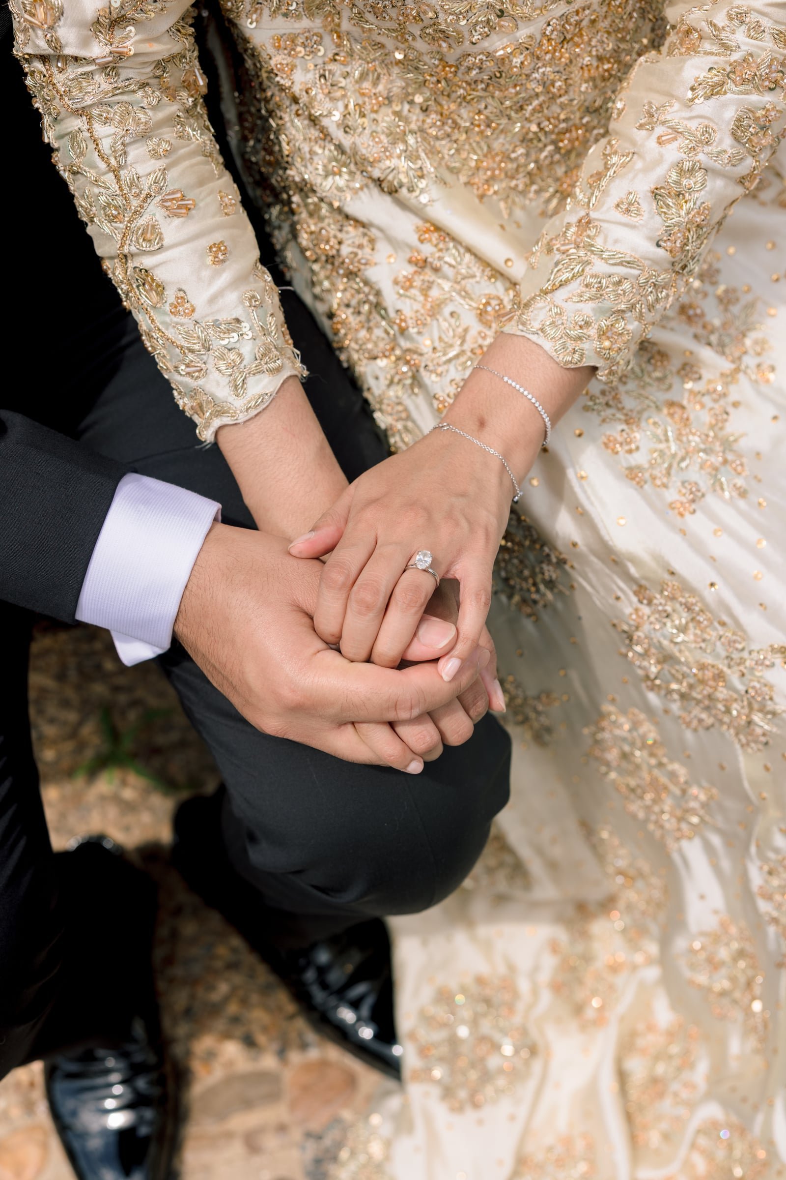 Close-up of a couple holding hands, with the woman's hand displaying an engagement ring and a delicate bracelet. The woman is wearing a cream-colored, heavily embroidered gown with gold and pearl embellishments. The man is in a black tuxedo with a wh