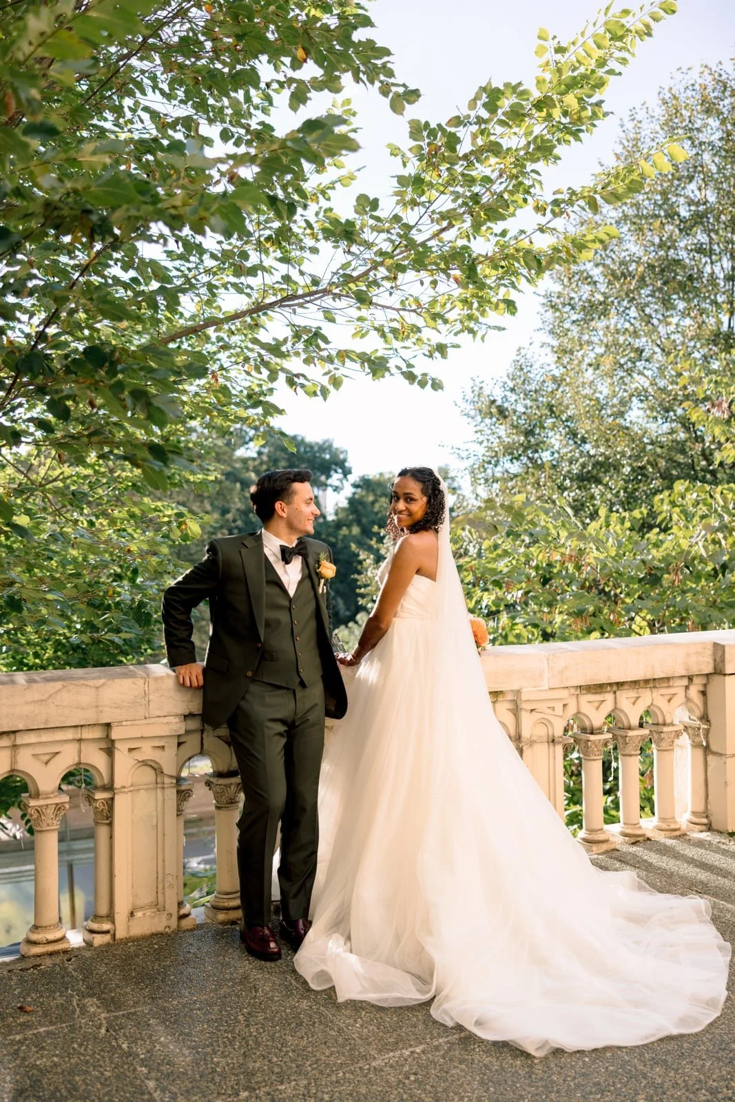 A bride and groom standing on a stone balcony with greenery and trees in the background, smiling and holding hands.
