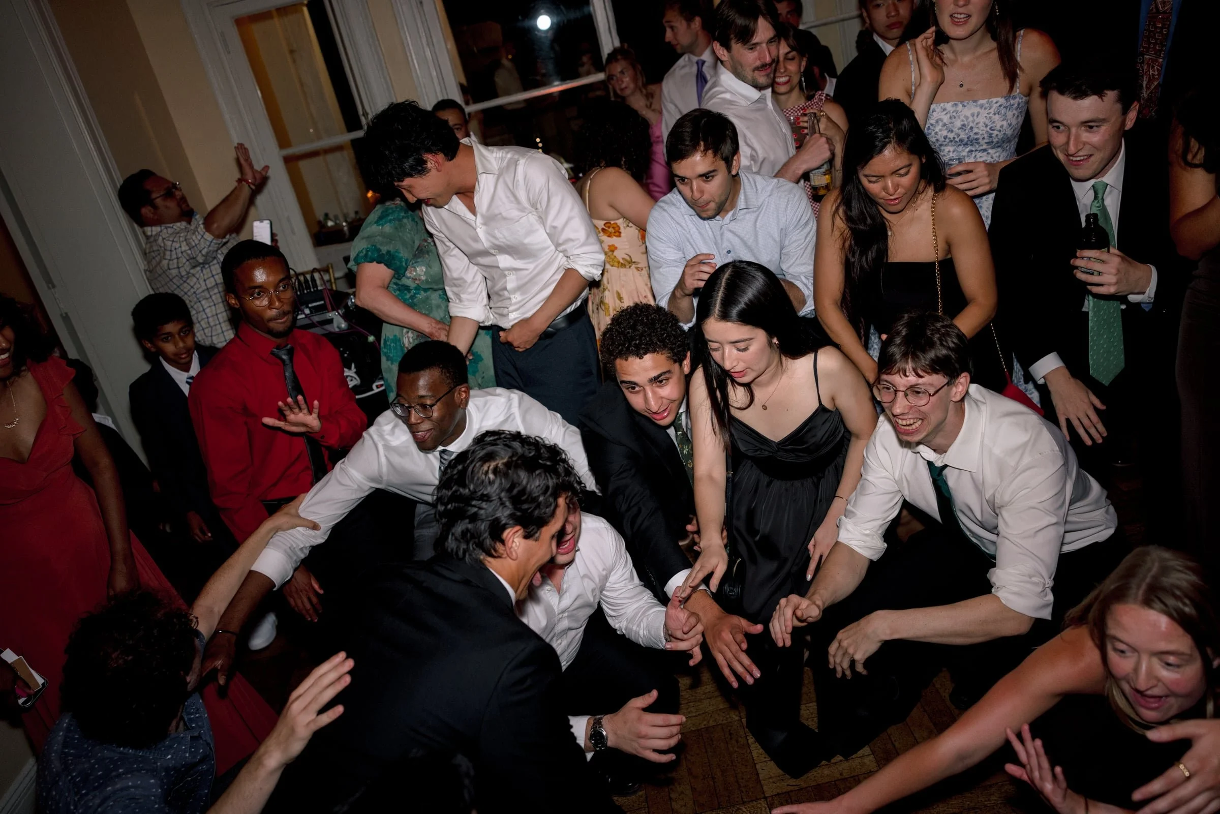Group of people dancing and socializing at a party, with some reaching out their hands and smiling, in a warmly lit indoor setting.
