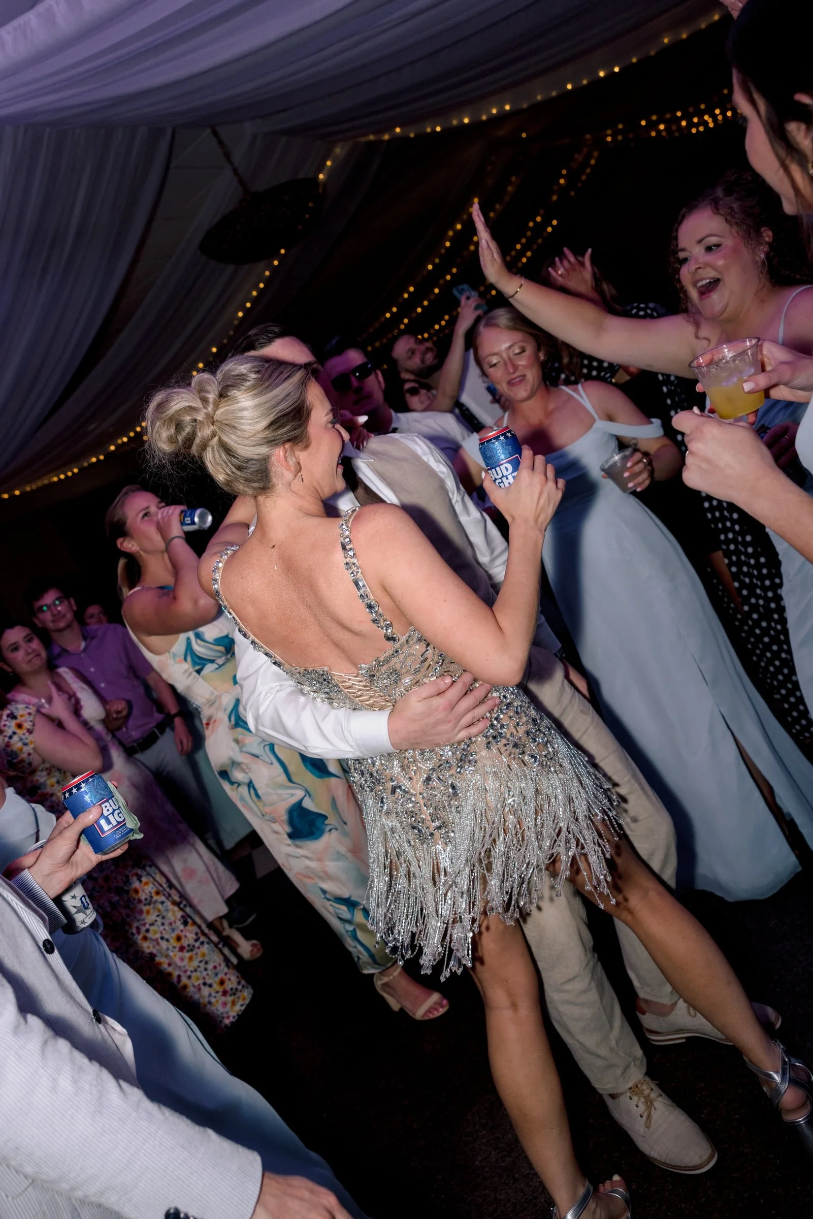People dancing and socializing at a lively party under a decorated ceiling with string lights, holding drinks and enjoying themselves.