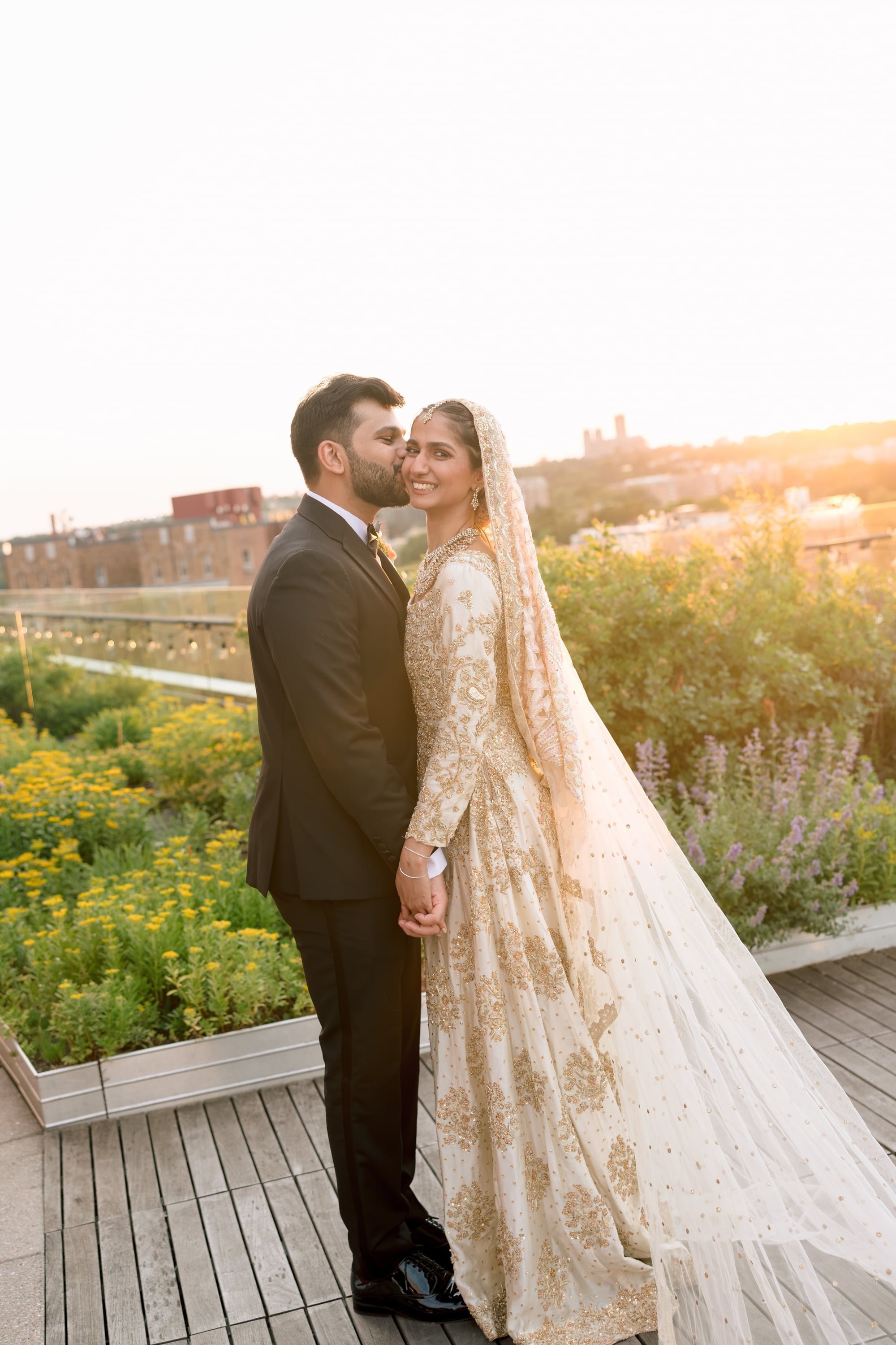 A bride and groom standing on a wooden deck holding hands, with the groom kissing the bride on her cheek during sunset, surrounded by flowers and city buildings in the background.