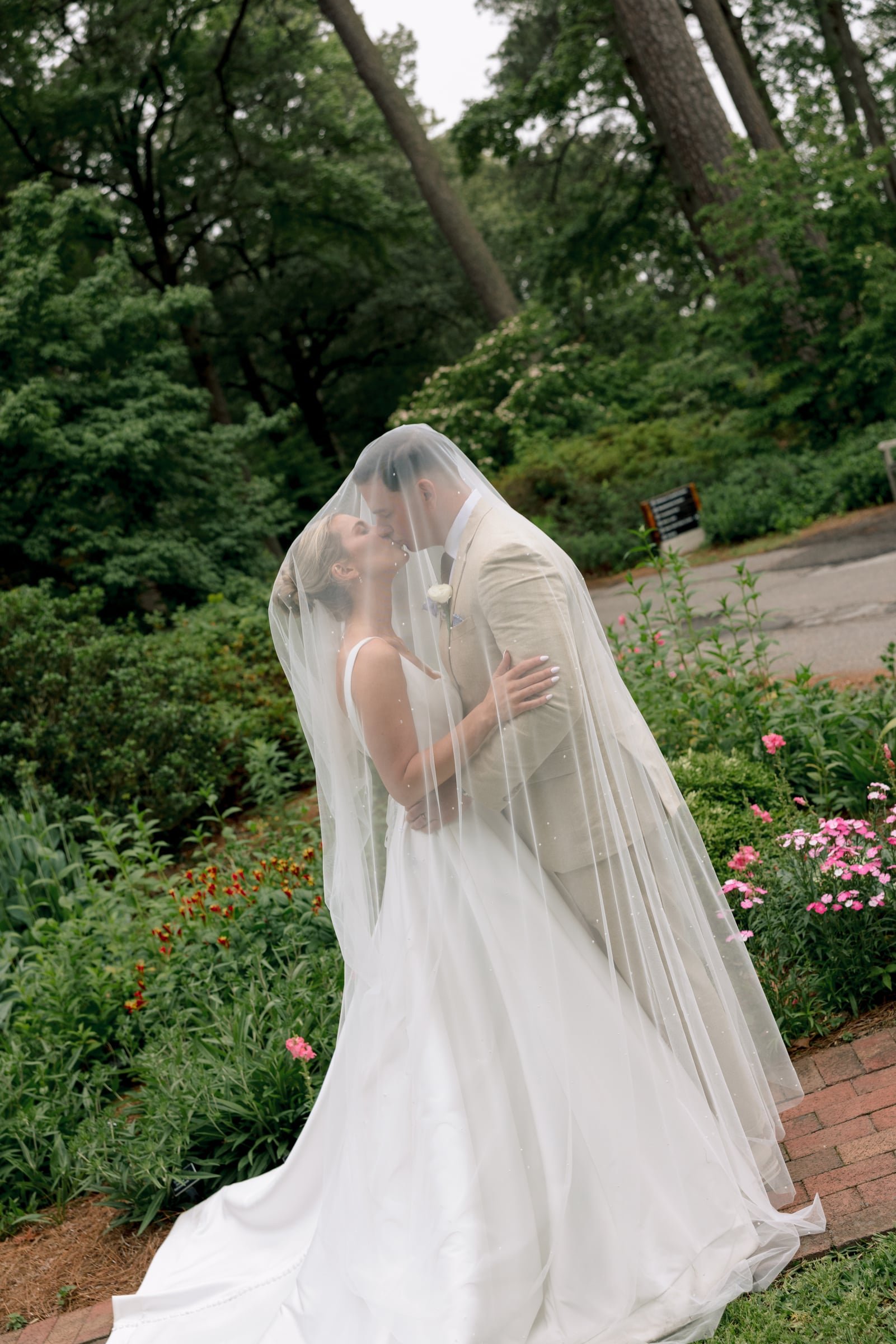 A bride and groom sharing a kiss under a towel, standing on a garden path surrounded by greenery and pink flowers.