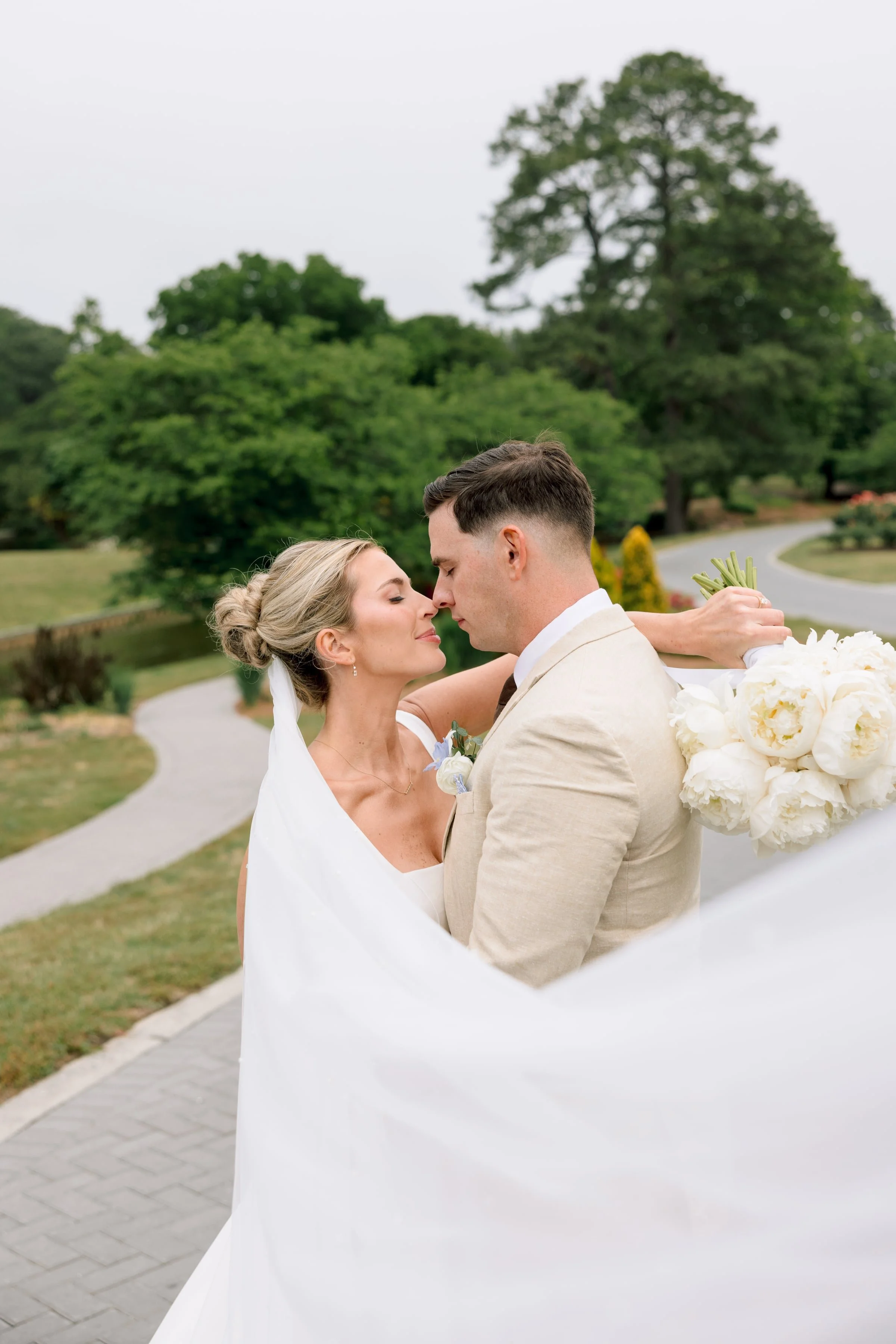 A bride and groom are embracing outdoors during their wedding, with the bride holding a bouquet of white flowers and the groom in a light-colored suit, with large trees and a curved path in the background.